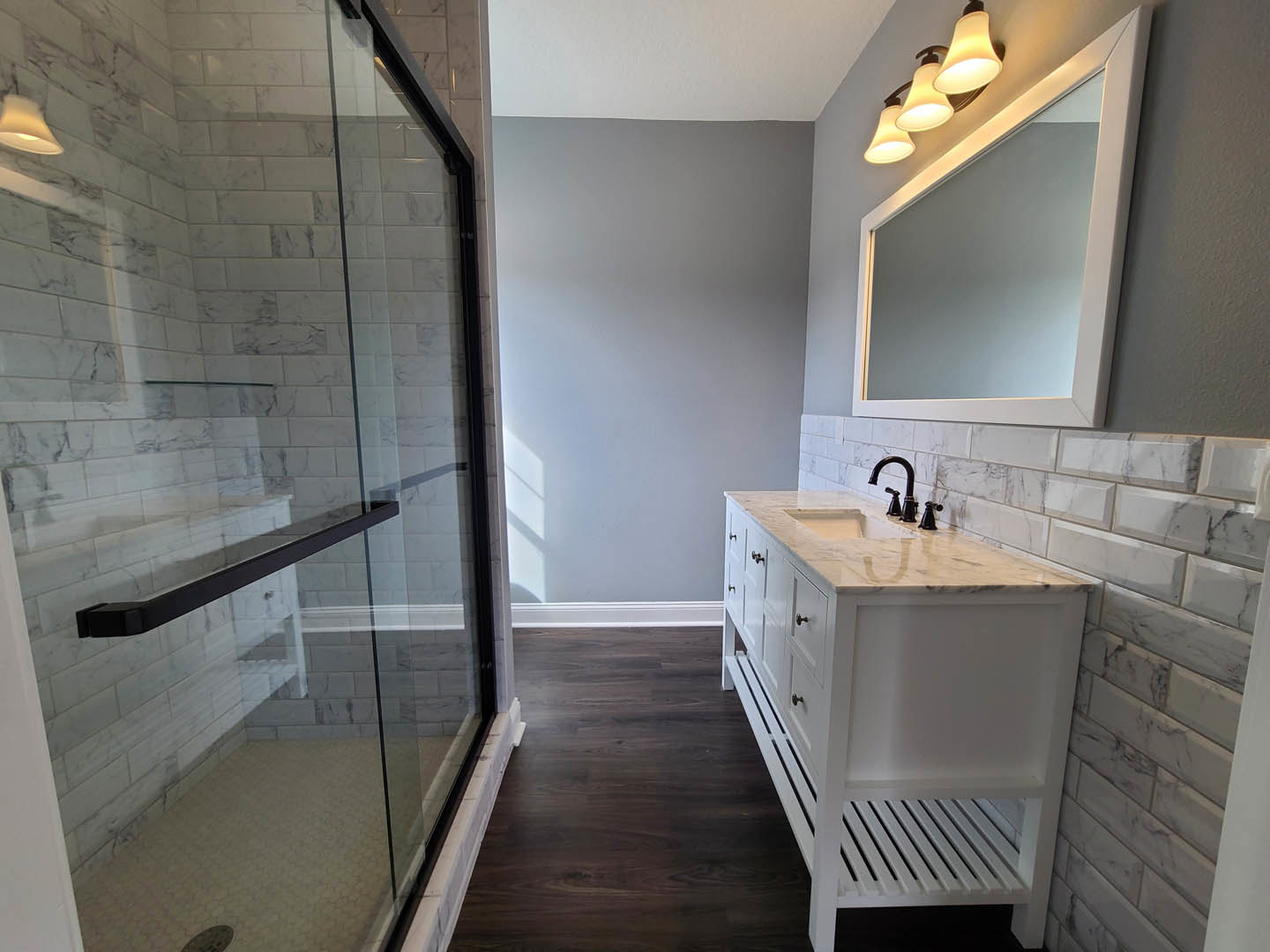 White bathroom vanity with marble countertop, black faucet, rectangular mirror with overhead light fixture, glass shower door with black hardware, white subway tile wall.