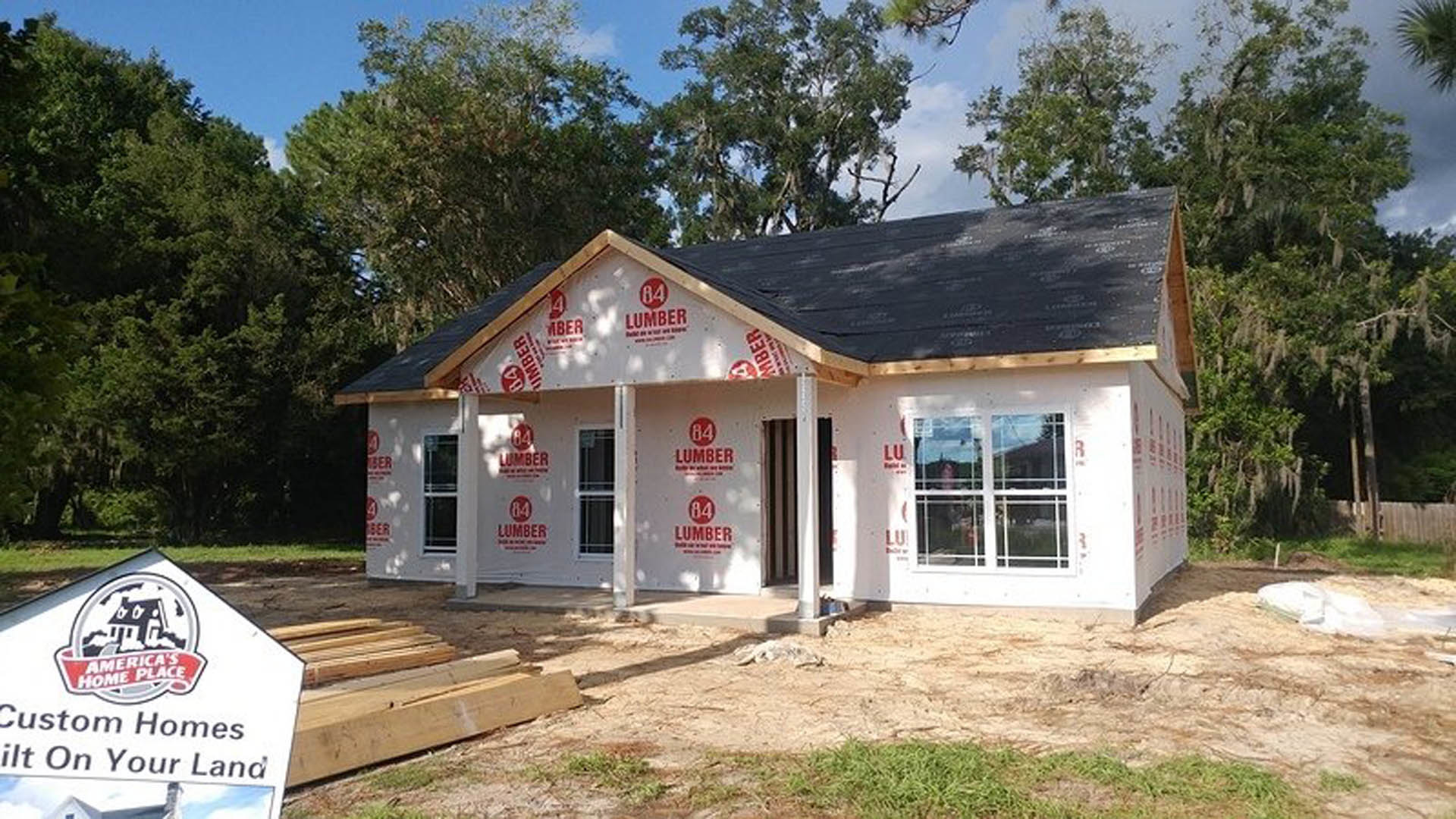 Framed house under construction with exposed lumber, white roof, white-framed window, construction sign in foreground, surrounded by trees and sky