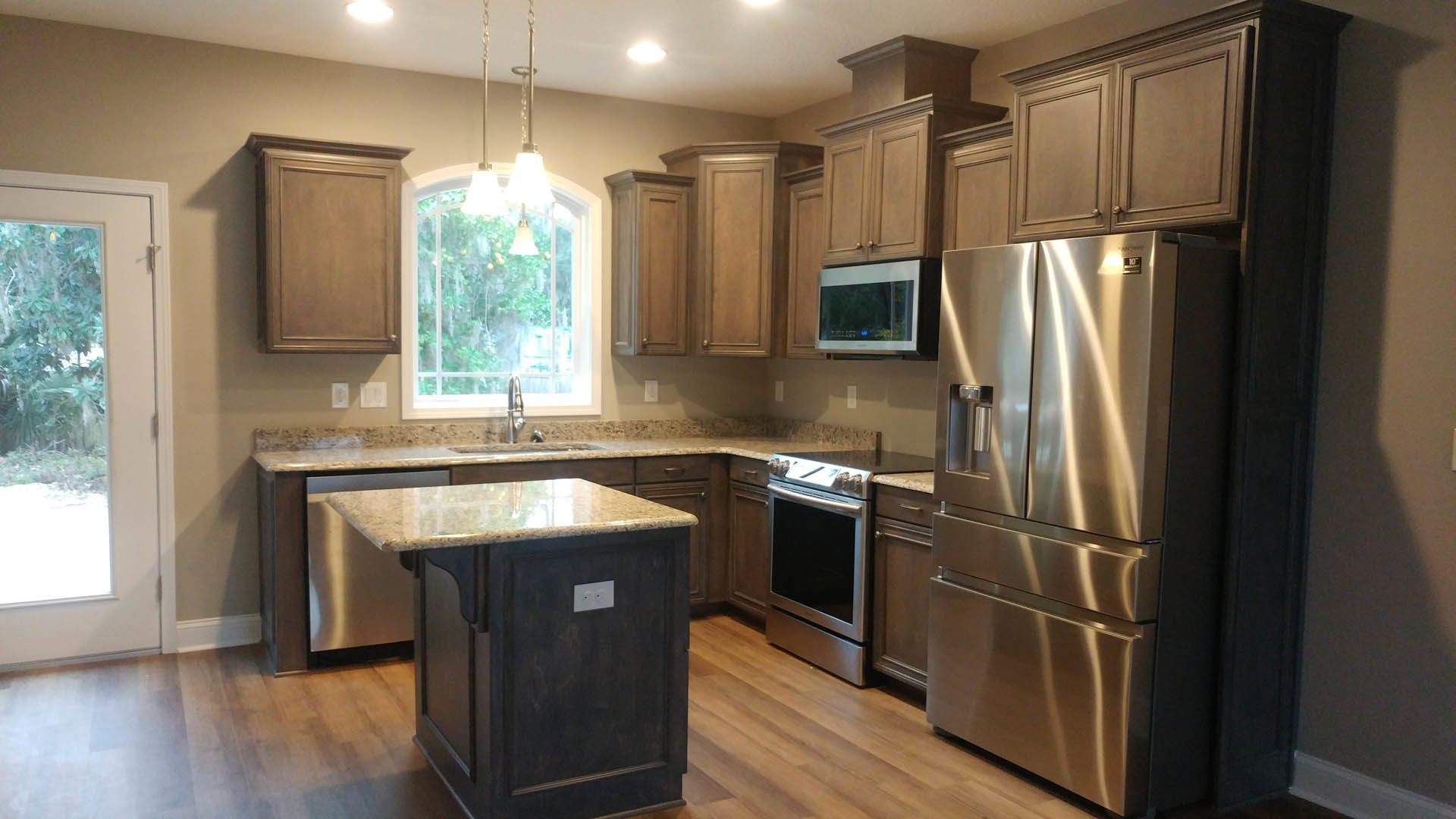Kitchen with stainless steel refrigerator, microwave, and sink; white cabinetry, stone countertops, window overlooking trees, and modern light fixture above sink.