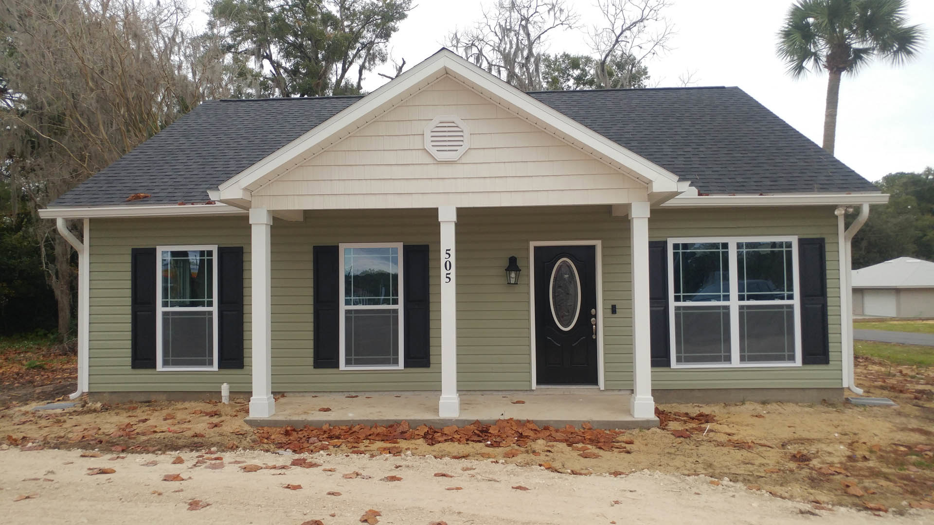 Black front door with white trim and column, white siding, grid window, sidewalk leading to entrance, small white house visible in background.