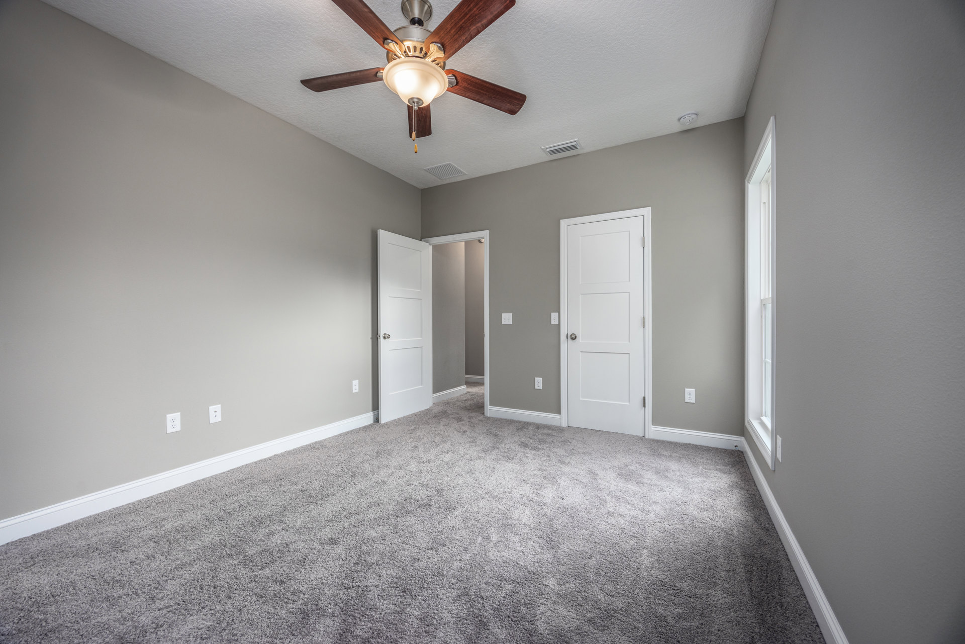 Carpeted room with white walls, ceiling fan with light fixture, two white doors with silver knobs, and white window frame