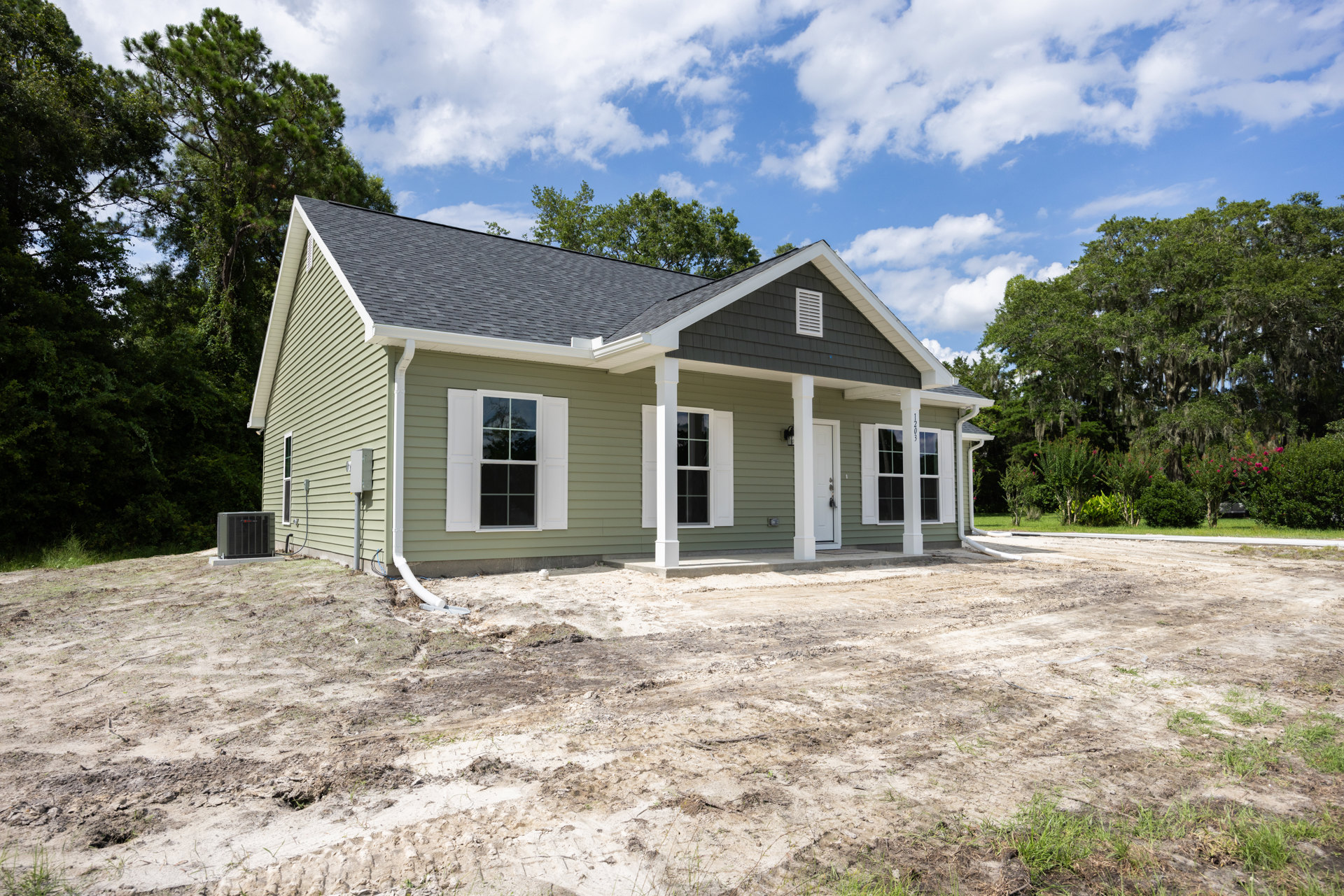 Green siding house with white shuttered windows, dirt lot in front, large leafy tree, and small white cottage in the background under cloudy sky.