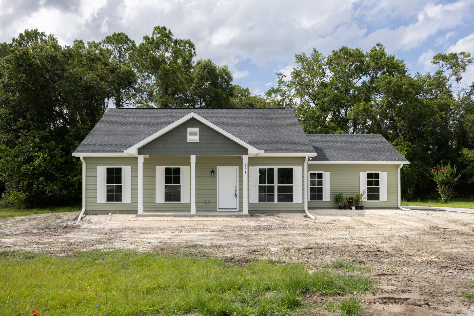 Two-story house surrounded by dirt and mature trees, white door with lock, multiple white-trimmed windows with shutters, grassy patches near dirt driveway, small white cottage