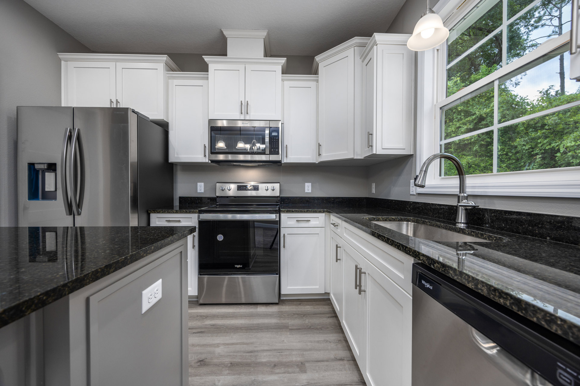 White kitchen cabinets with silver handles, black countertops, stainless steel oven and microwave, single pendant light fixture, window overlooking trees
