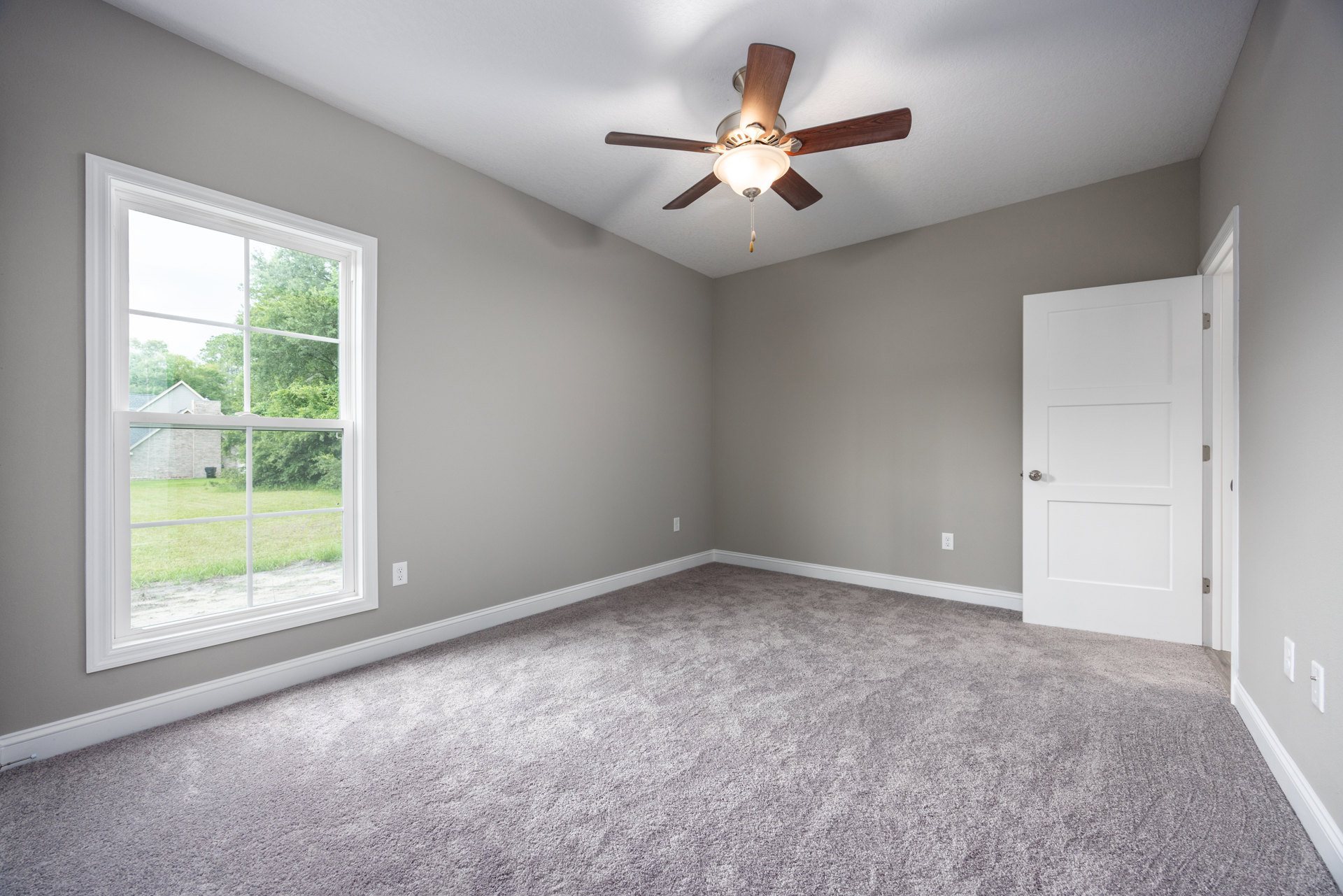 Carpeted room with white walls, ceiling fan with light fixture, window overlooking yard and trees, white door with silver knob