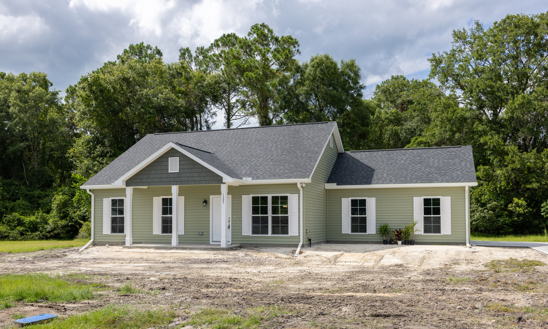 Partially built house with green roof, white-framed windows, and white shutters, surrounded by trees; dirt patch in front, small white cottage visible in background.