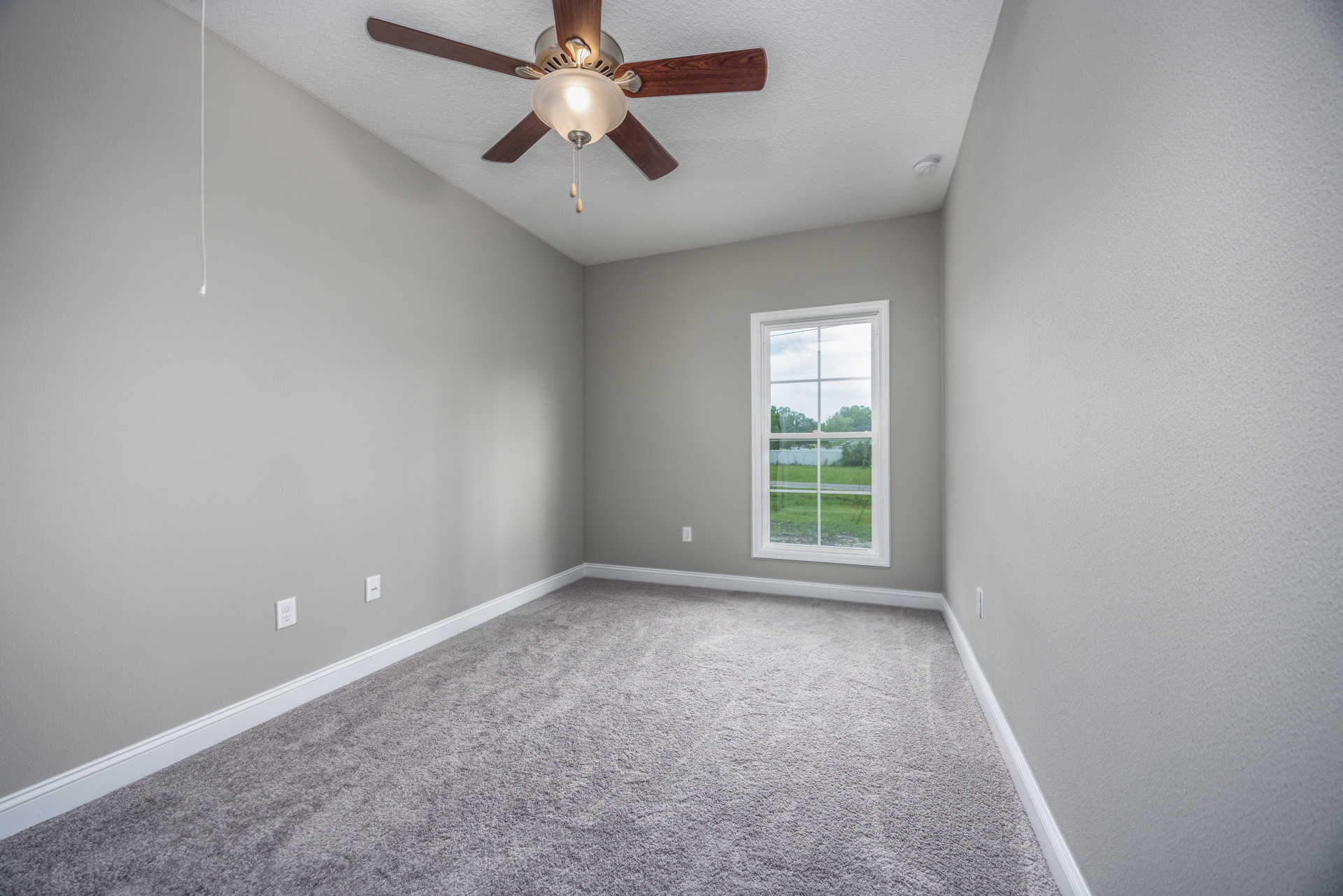 Carpeted room with white plaster walls, ceiling fan with light fixture, large window overlooking green yard and trees
