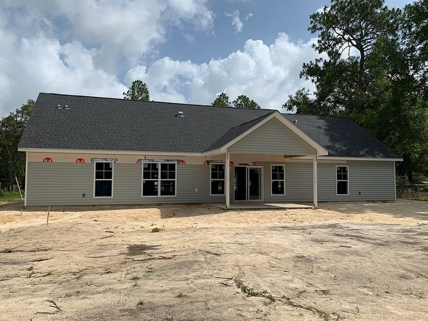 Two-story home under construction with exposed wood framing, white window frames, covered porch, dirt yard, and surrounding trees