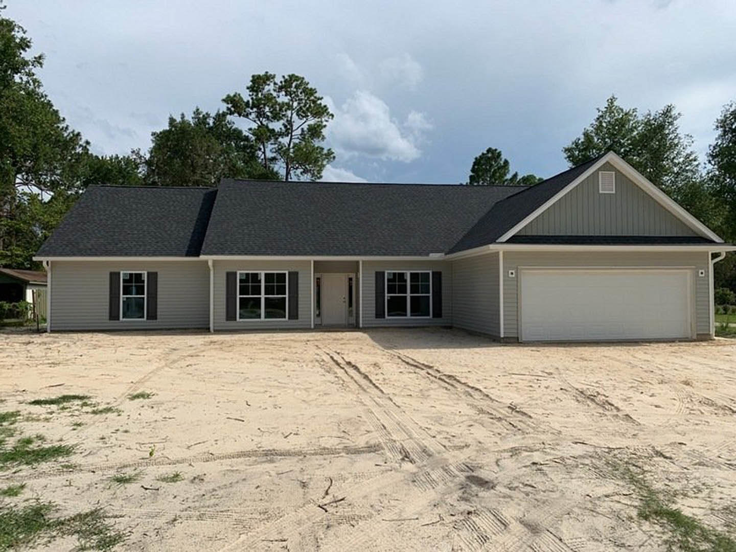 White modern house with black roof, white-framed windows, and glass-paneled front door; sandy driveway with visible tire tracks; bordered by trees under a cloudy sky.