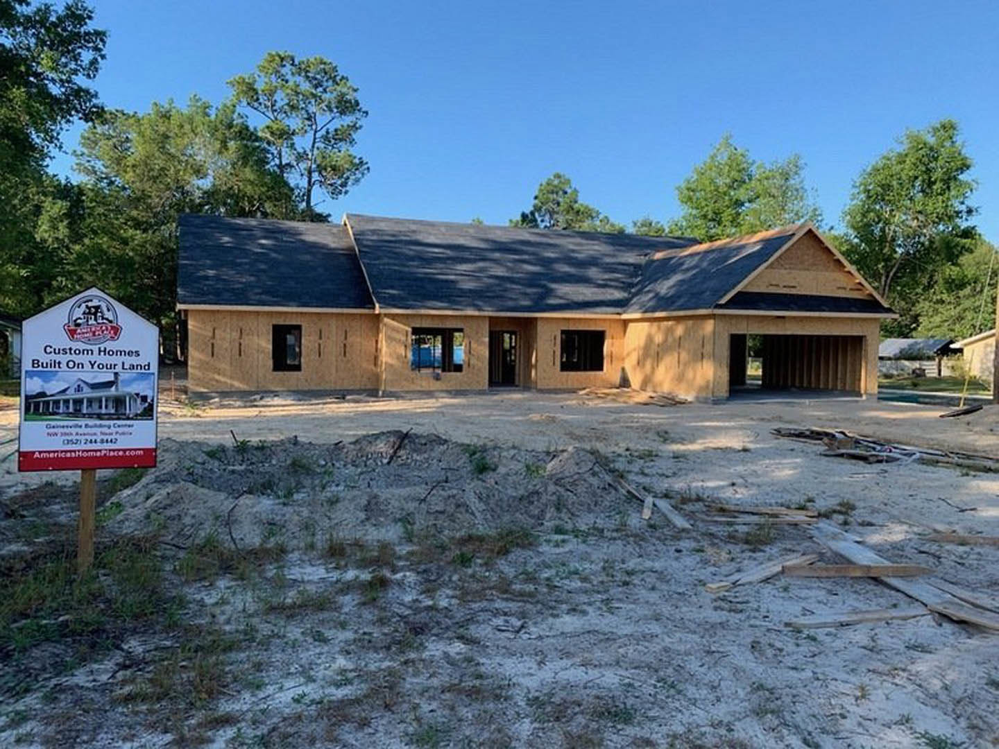 Wood-framed house under construction with exposed sheathing, surrounded by trees, construction sign displaying house rendering in front yard