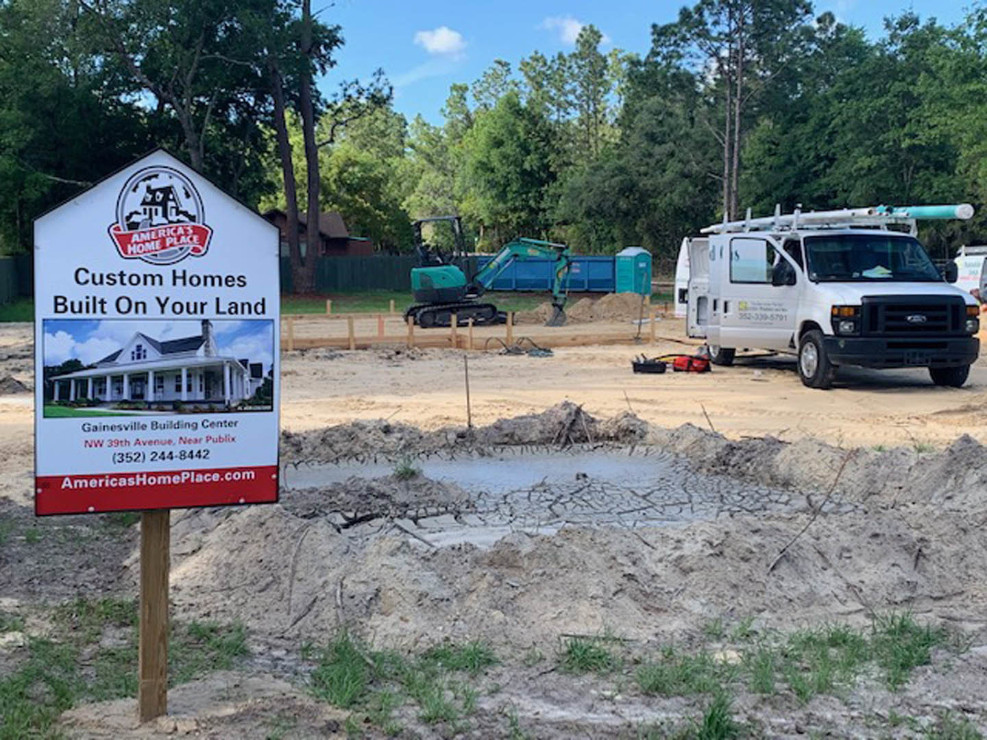 Construction site with dirt ground, white van with roof ladder parked beside a temporary sign, trees and blue sky in background