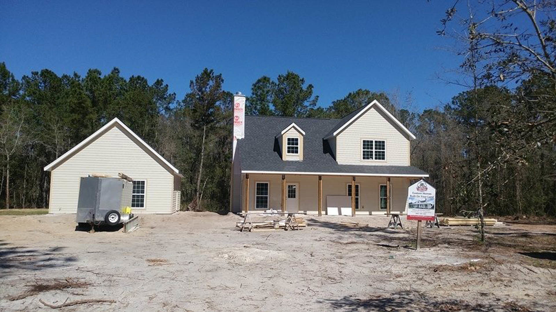Wood-framed house under construction in a wooded area with a covered porch, white-framed windows, and a utility trailer parked in front