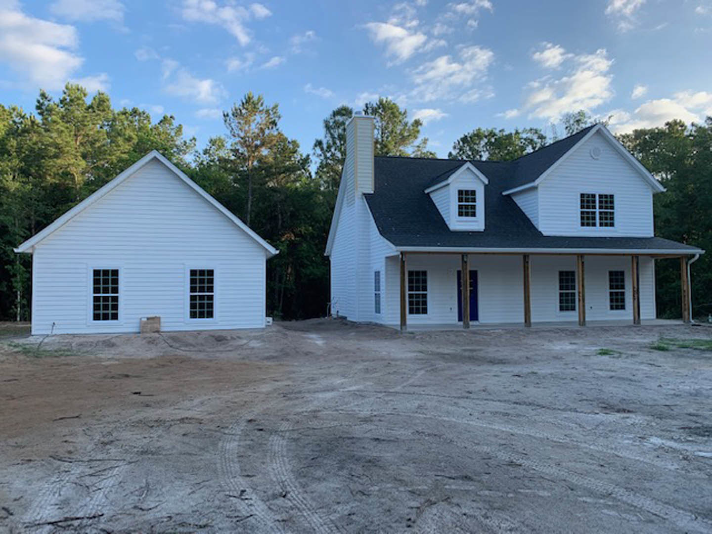 White house with blue door, grid-style windows, tire tracks on dirt yard, surrounded by trees under cloudy sky