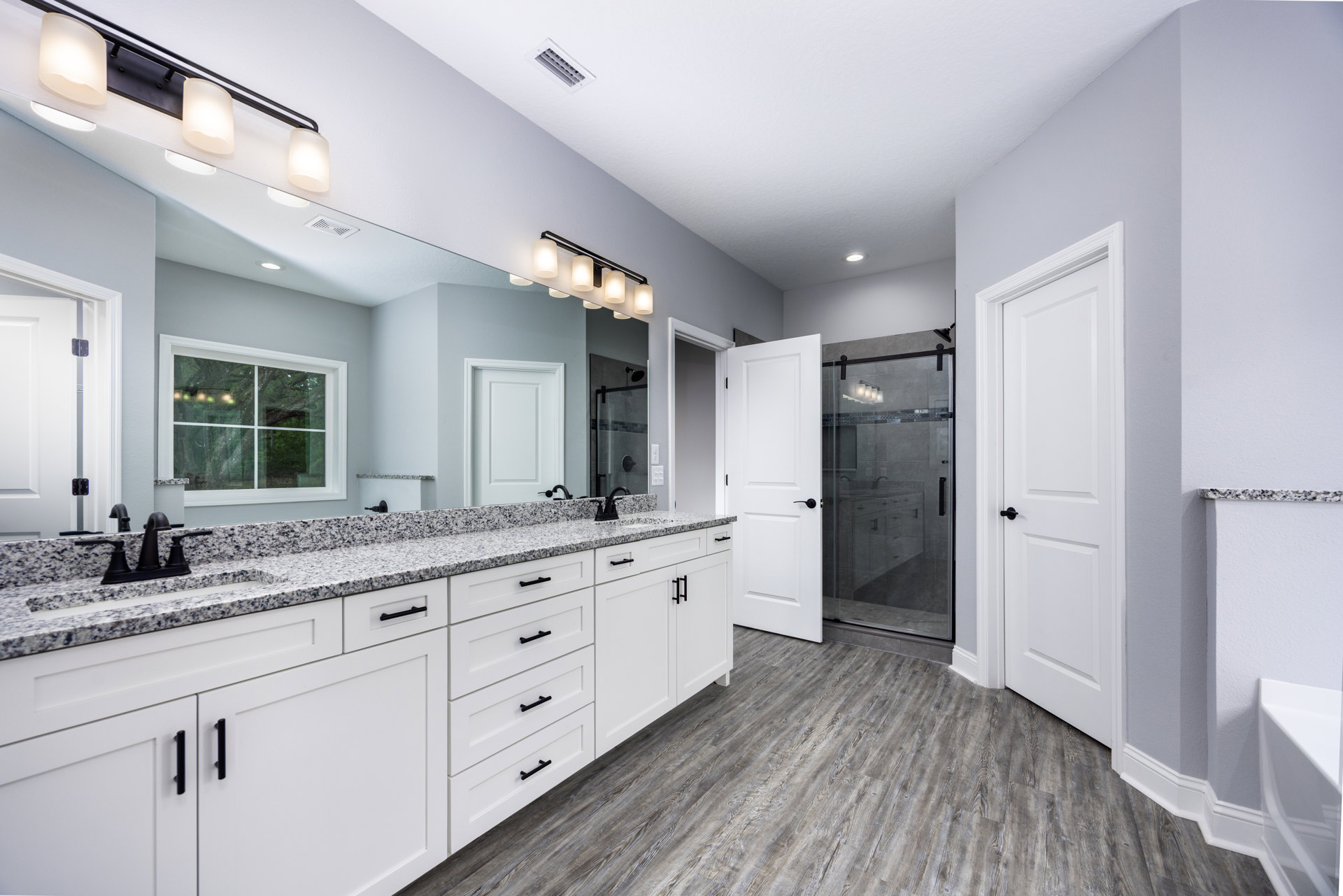 Bathroom with white shaker cabinets, wide frameless mirror above quartz countertop, chrome faucet, glass shower door, white paneled door with black handle, window with white trim