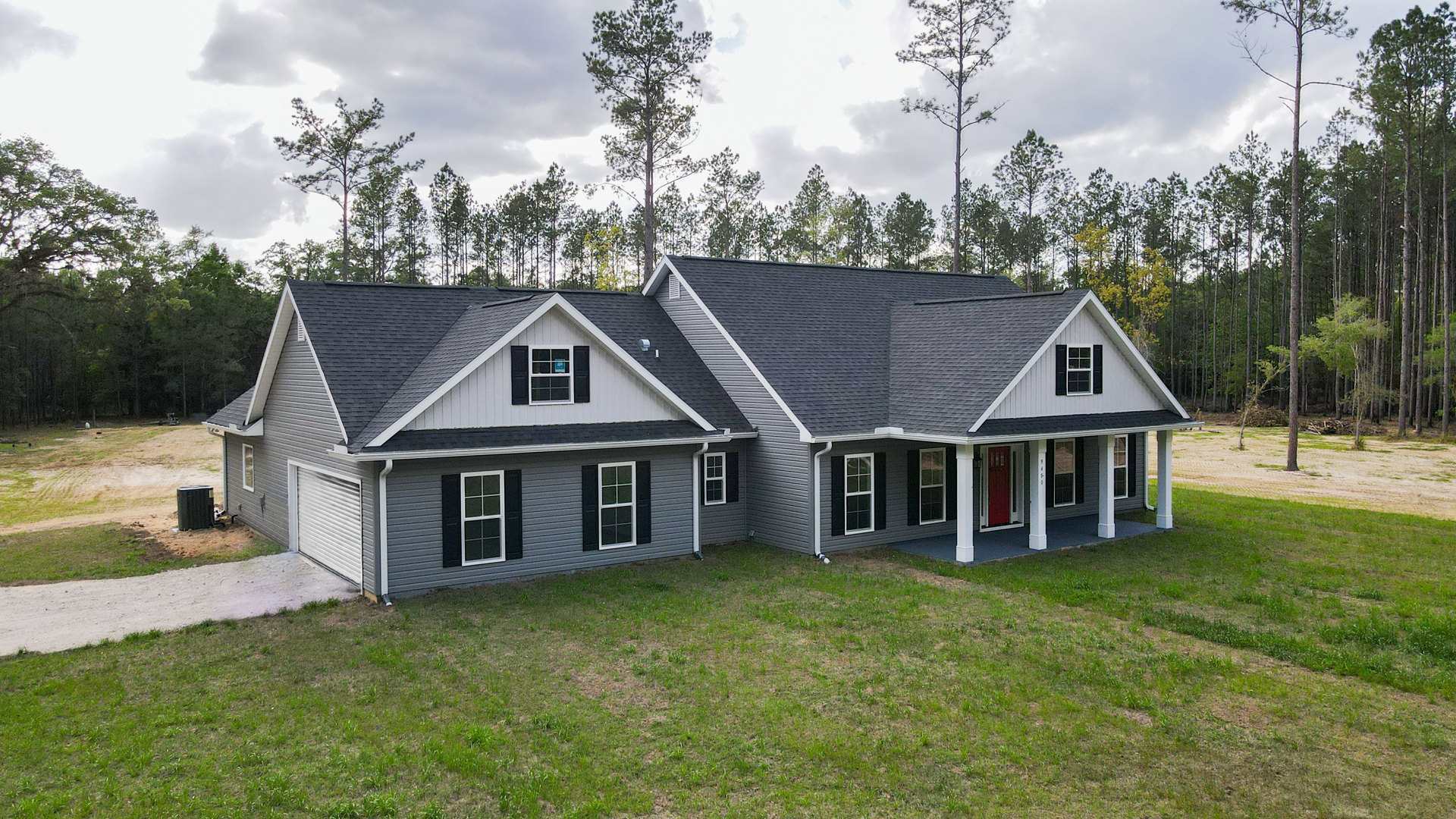 Two-story house with gray siding, white trim, large windows, manicured green lawn, mature trees in background, cloudy sky overhead