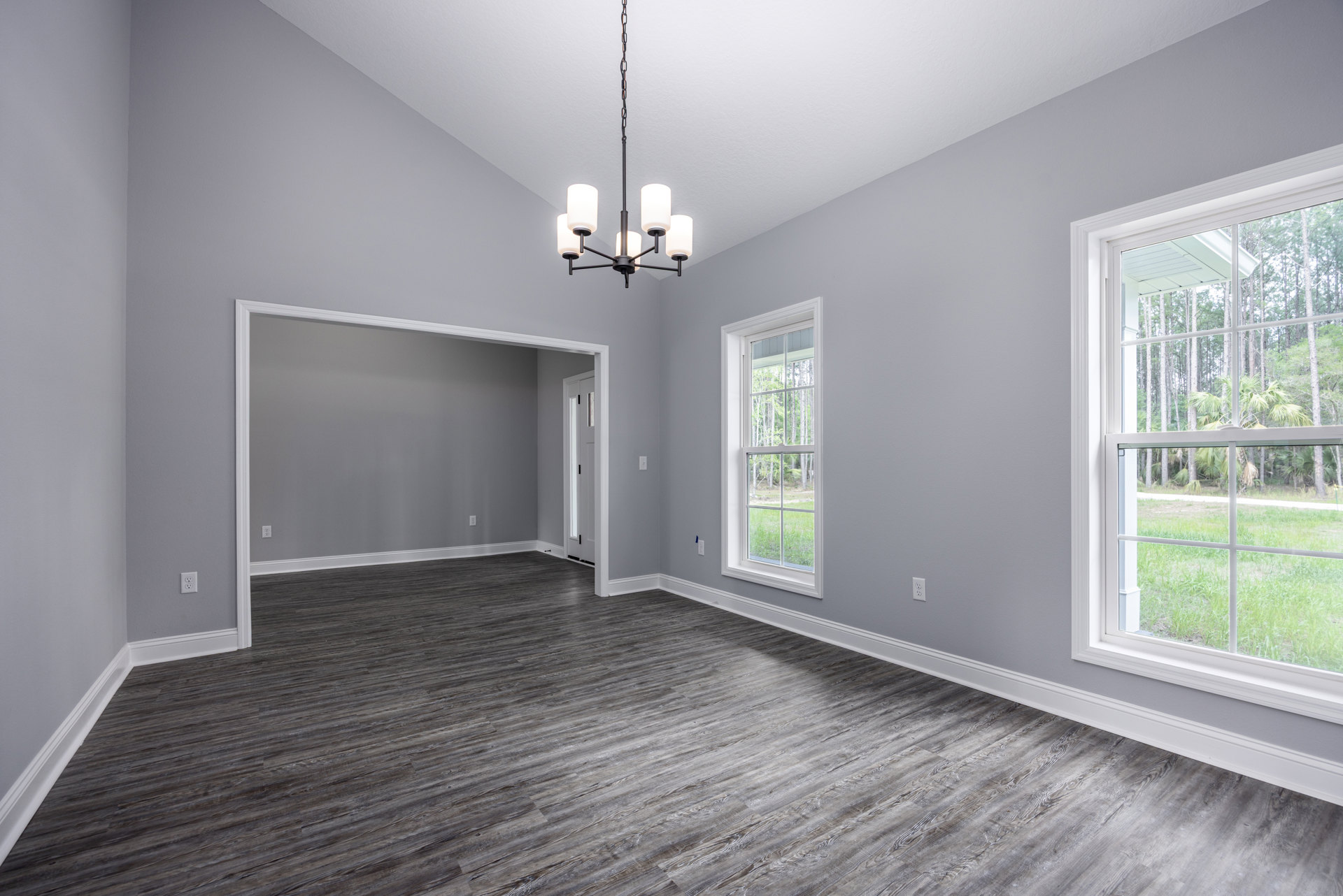 Wood flooring in a spacious room with a large window overlooking trees and grass, white plaster walls, and a ceiling-mounted chandelier with white lamps.