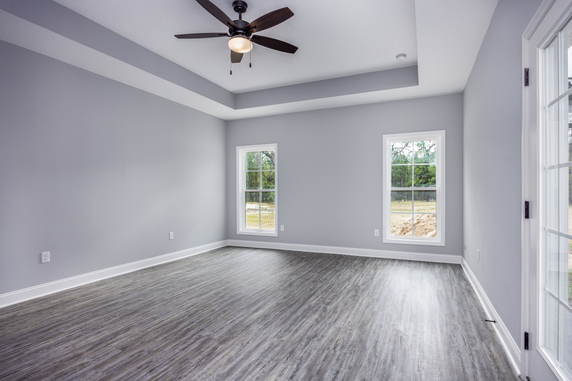 Ceiling fan with light fixture, multiple windows showing trees outside, wood flooring, white plaster walls, black window hinges