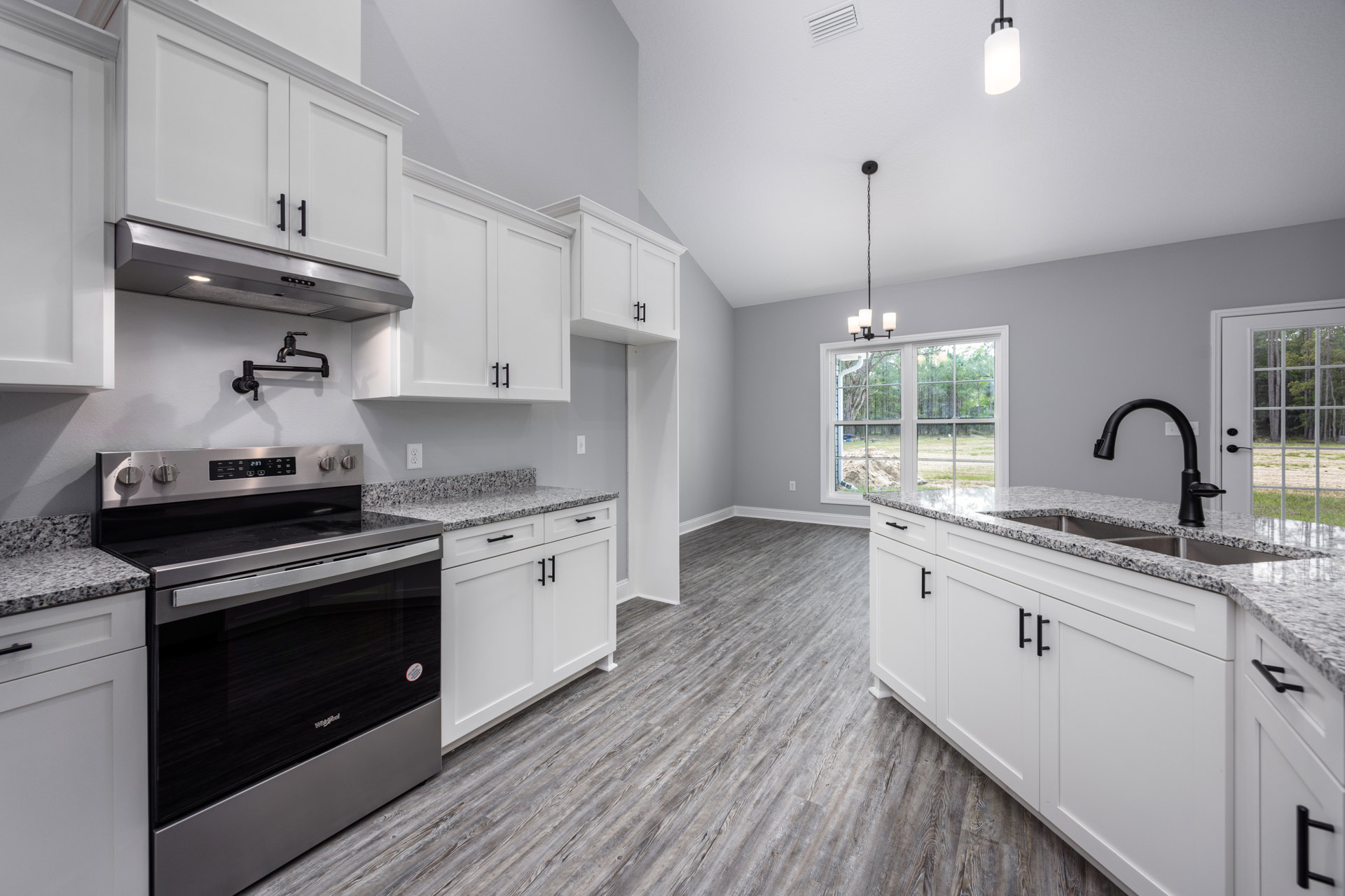 White shaker cabinets, wood plank flooring, black glass stove, white kitchen island with black legs, black faucet on quartz countertop, glass door revealing trees outside