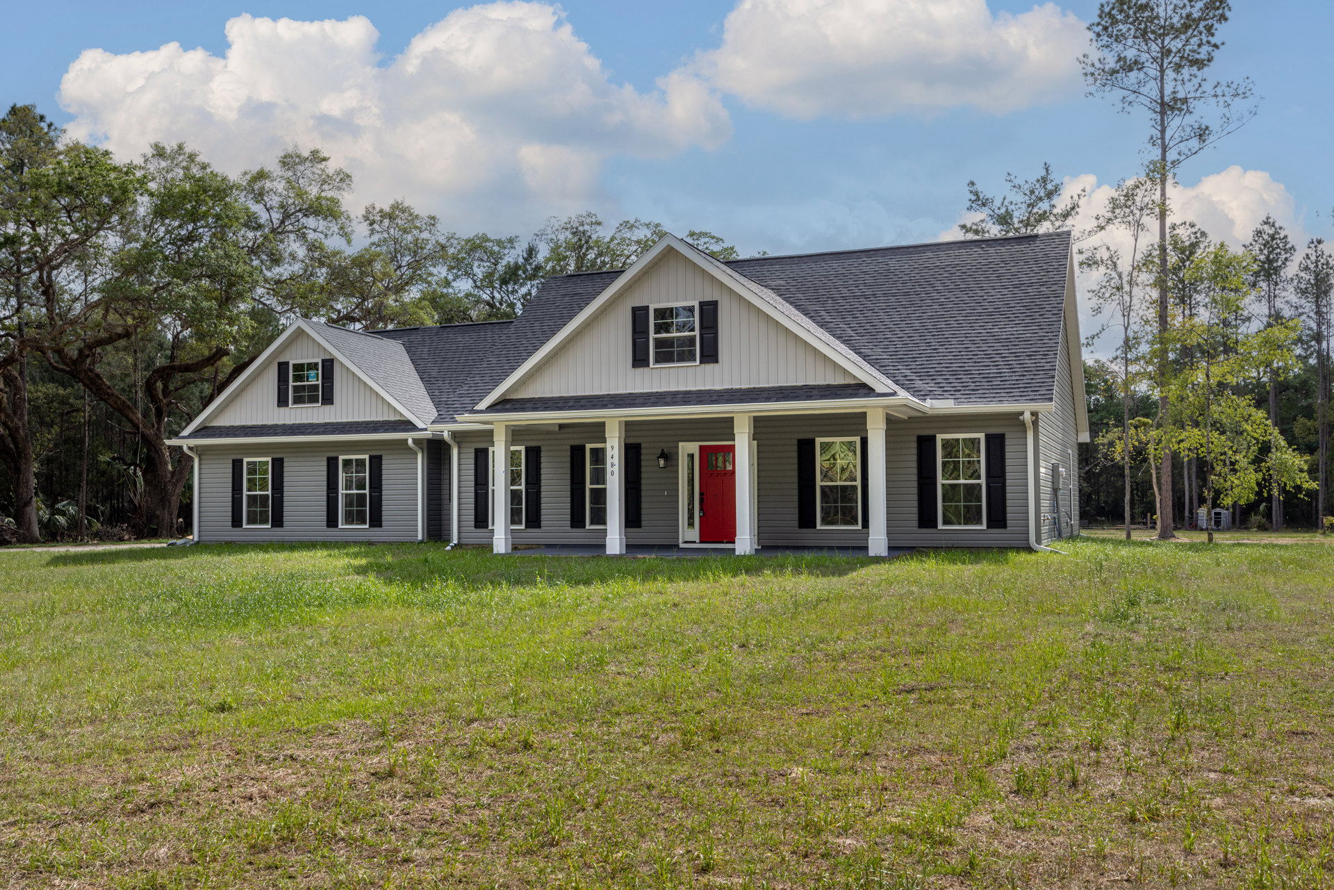 White siding house with a red front door, covered porch, green lawn, and mature trees in the background