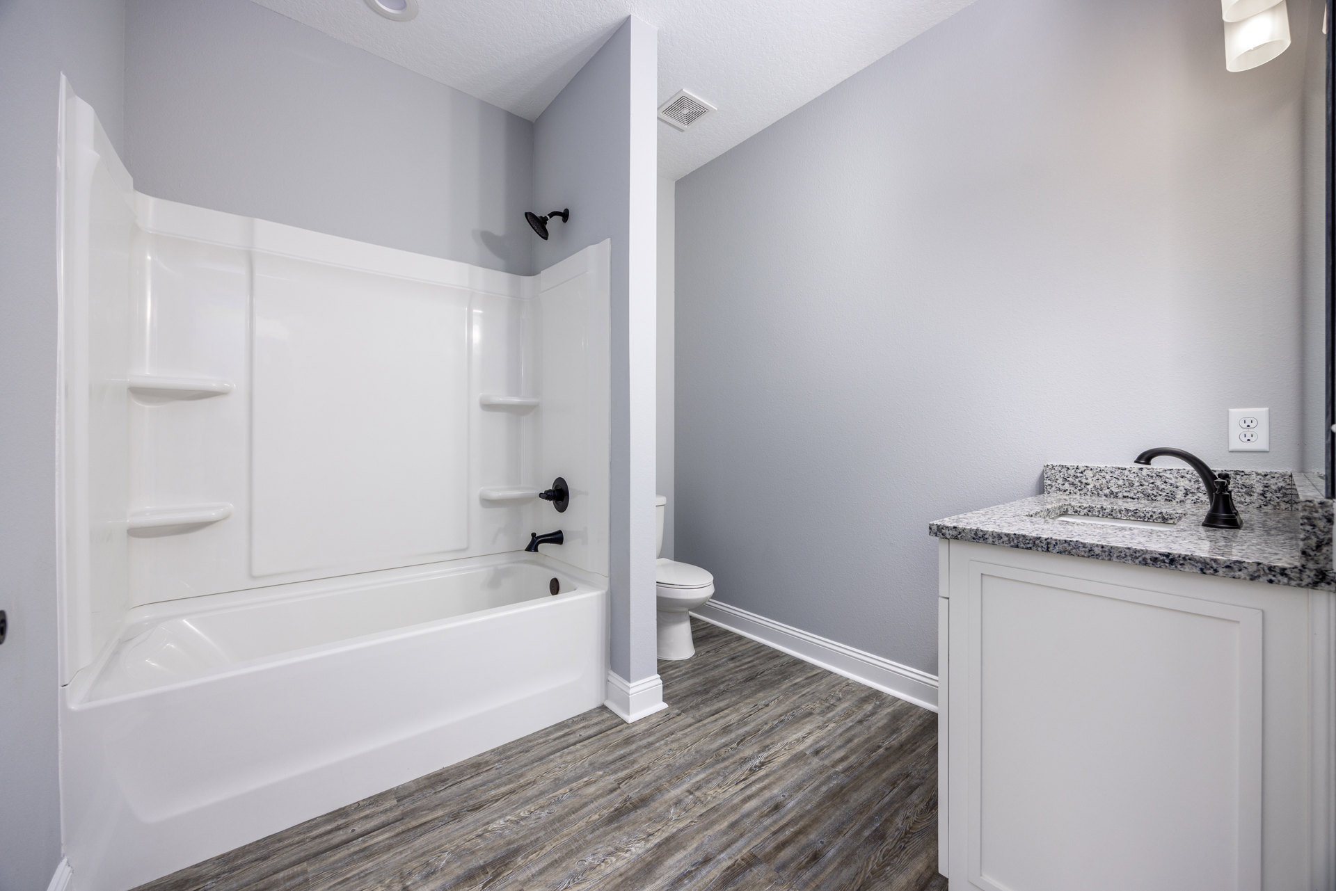 White bathtub and toilet in a bathroom with light tile flooring, wall-mounted shower head, ceiling vent, and electrical outlet visible near cabinetry