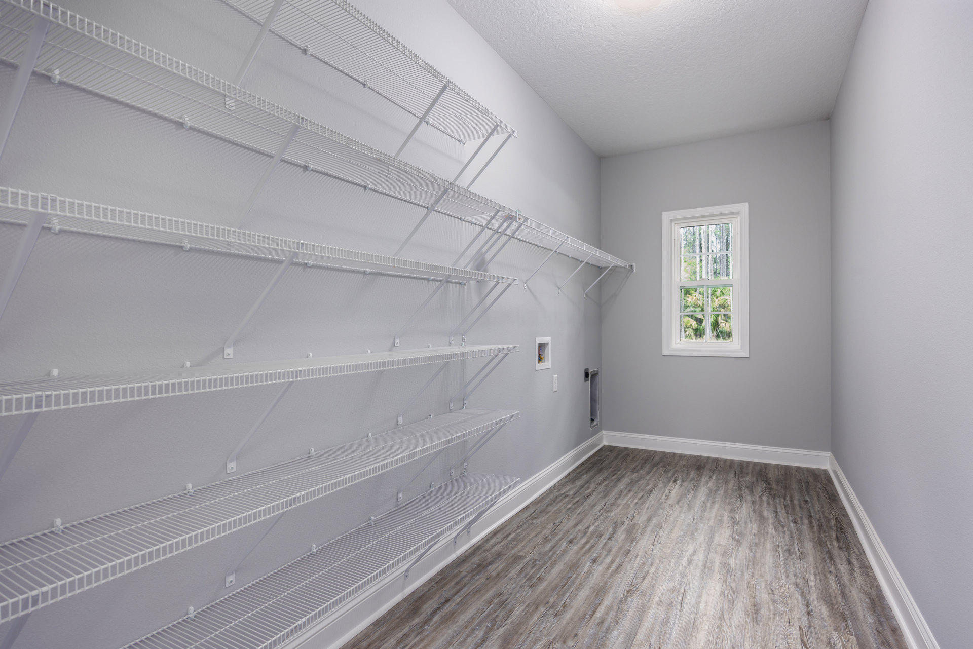 Spacious room featuring white wall shelves, light wood flooring with white trim, and a large window overlooking leafy trees.