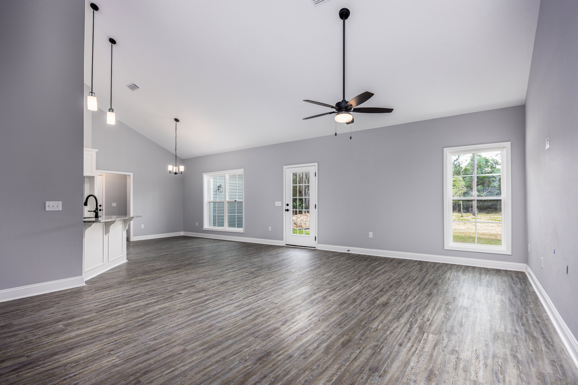 Spacious room with hardwood flooring, black-bladed ceiling fan, large window overlooking trees, plaster walls, and door opening to outdoor landscape.