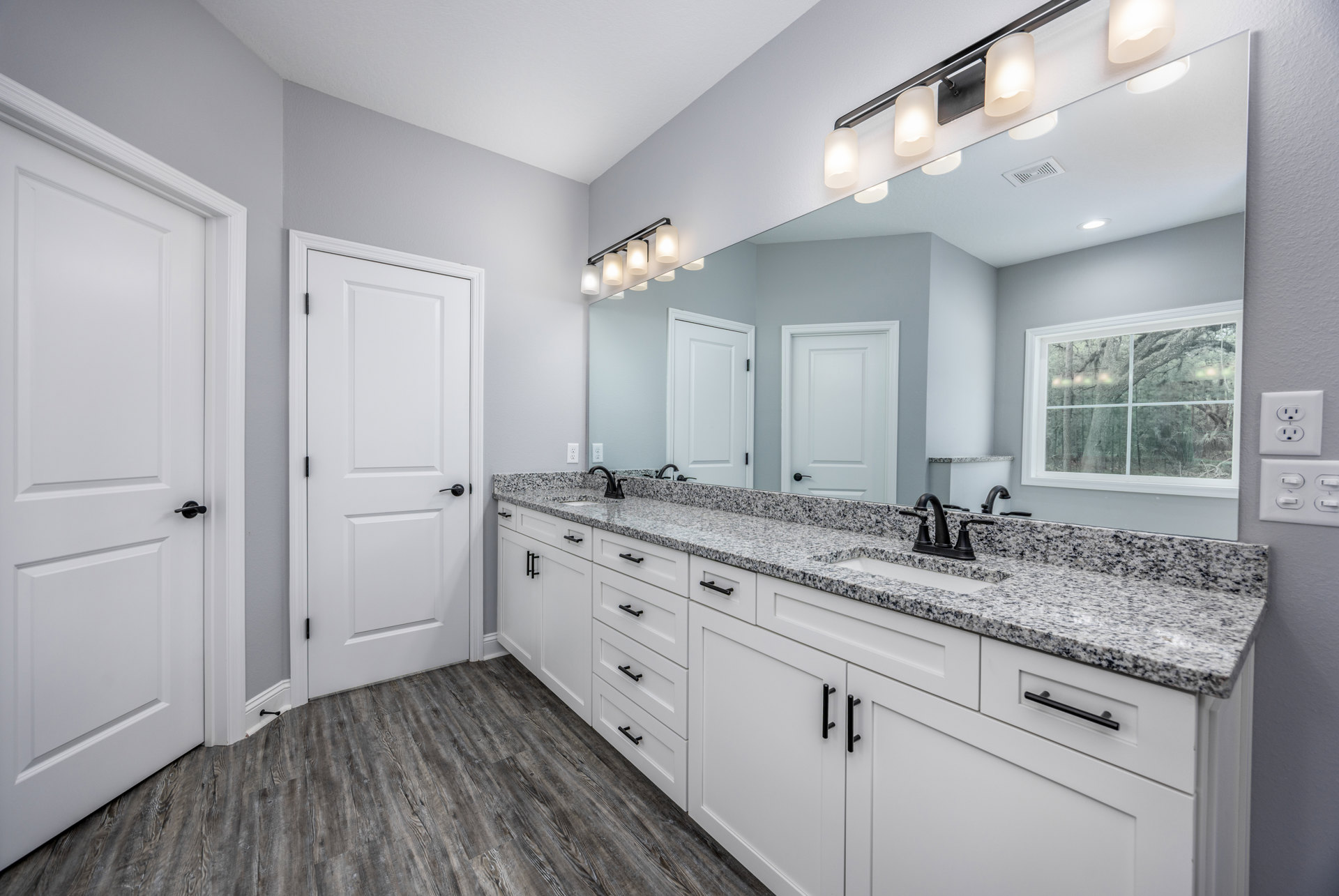 Bathroom featuring marble countertop, large rectangular mirror, white cabinetry, chrome faucet, white light fixture with shade, white door with black handle and hinge, window