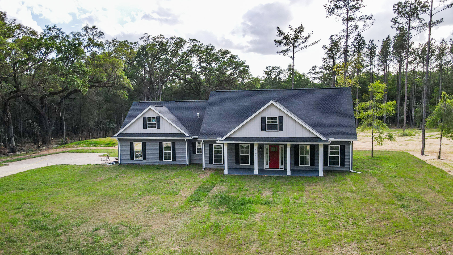 Two-story house with white siding, red front door with white trim, large front lawn, white-framed windows, covered porch, and mature trees under a partly cloudy sky