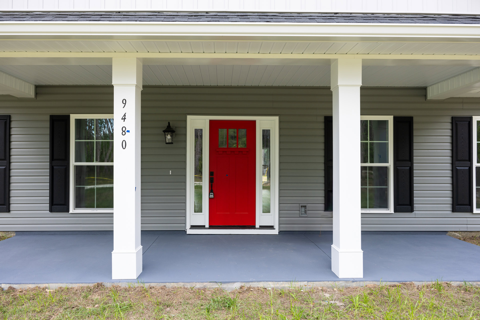 Red front door with silver lock, flanked by white pillars, set in light siding; adjacent window with white frame, grass and dirt beside concrete wall, white board with blue handle
