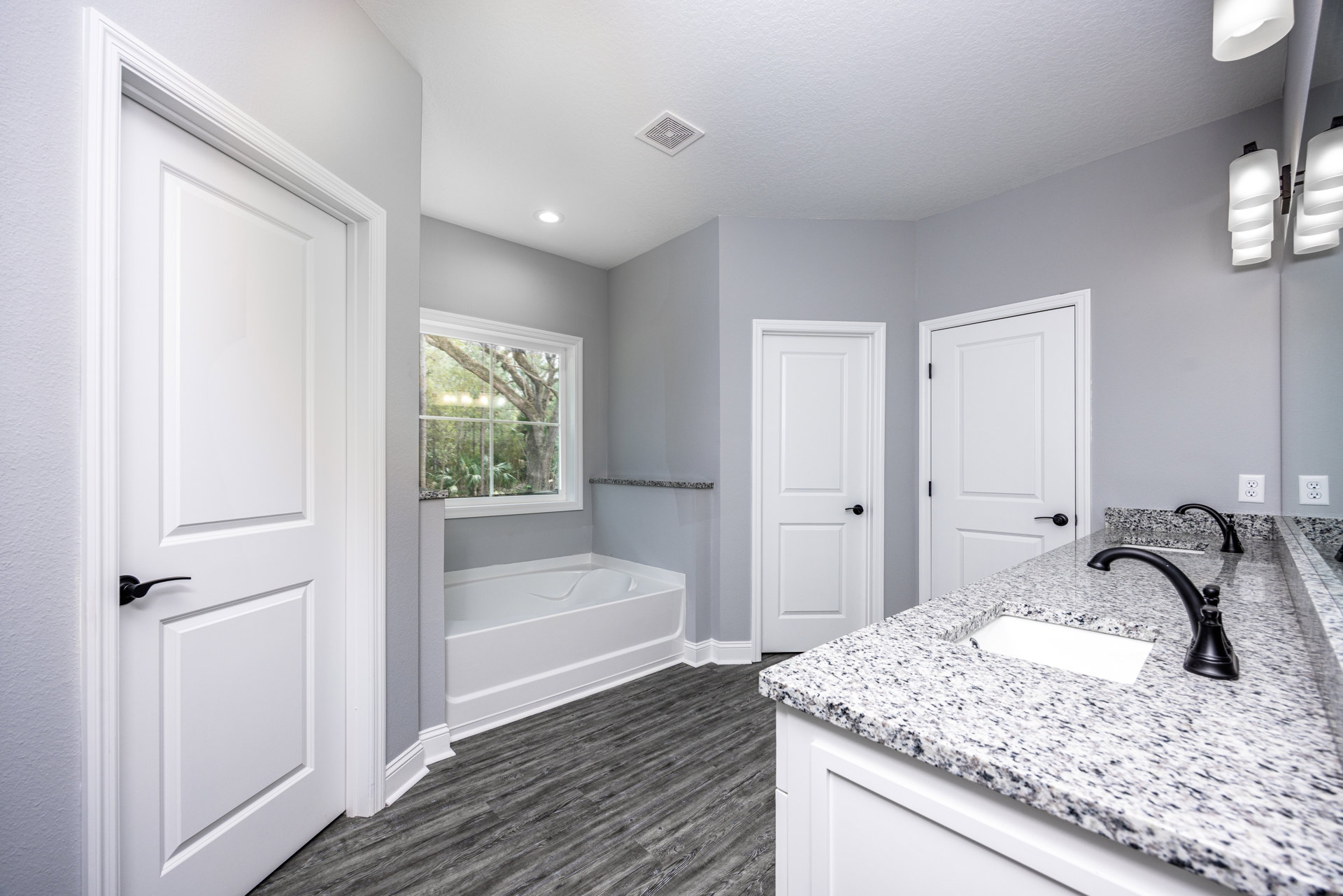 Freestanding bathtub beside a large window with trees visible outside, white vanity with undermount sink and chrome faucet, modern light fixture, white door with black handle