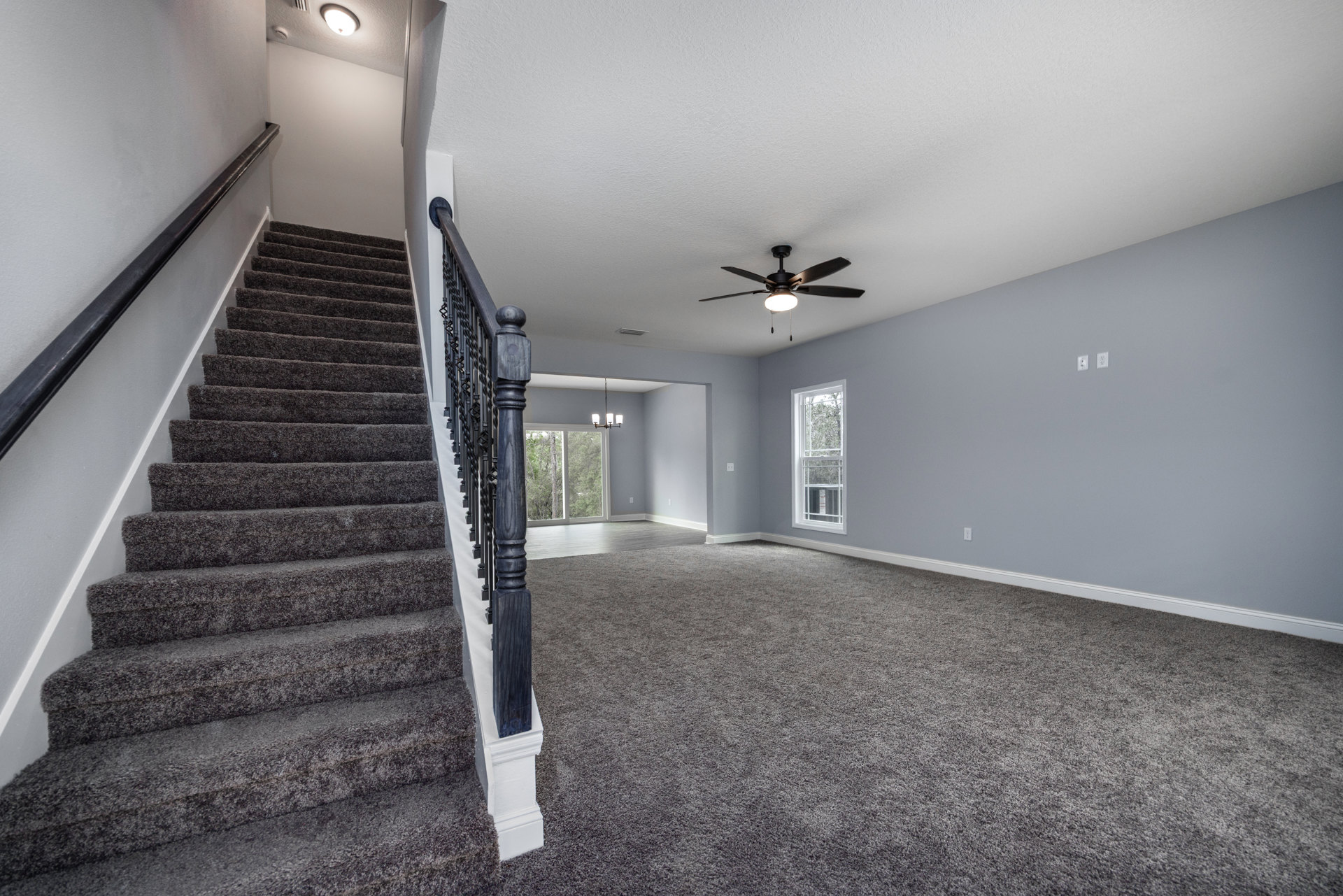 Wood staircase with white risers and handrail, ceiling fan with light above, large window framed in white showing green trees outside, plaster walls and hardwood flooring.