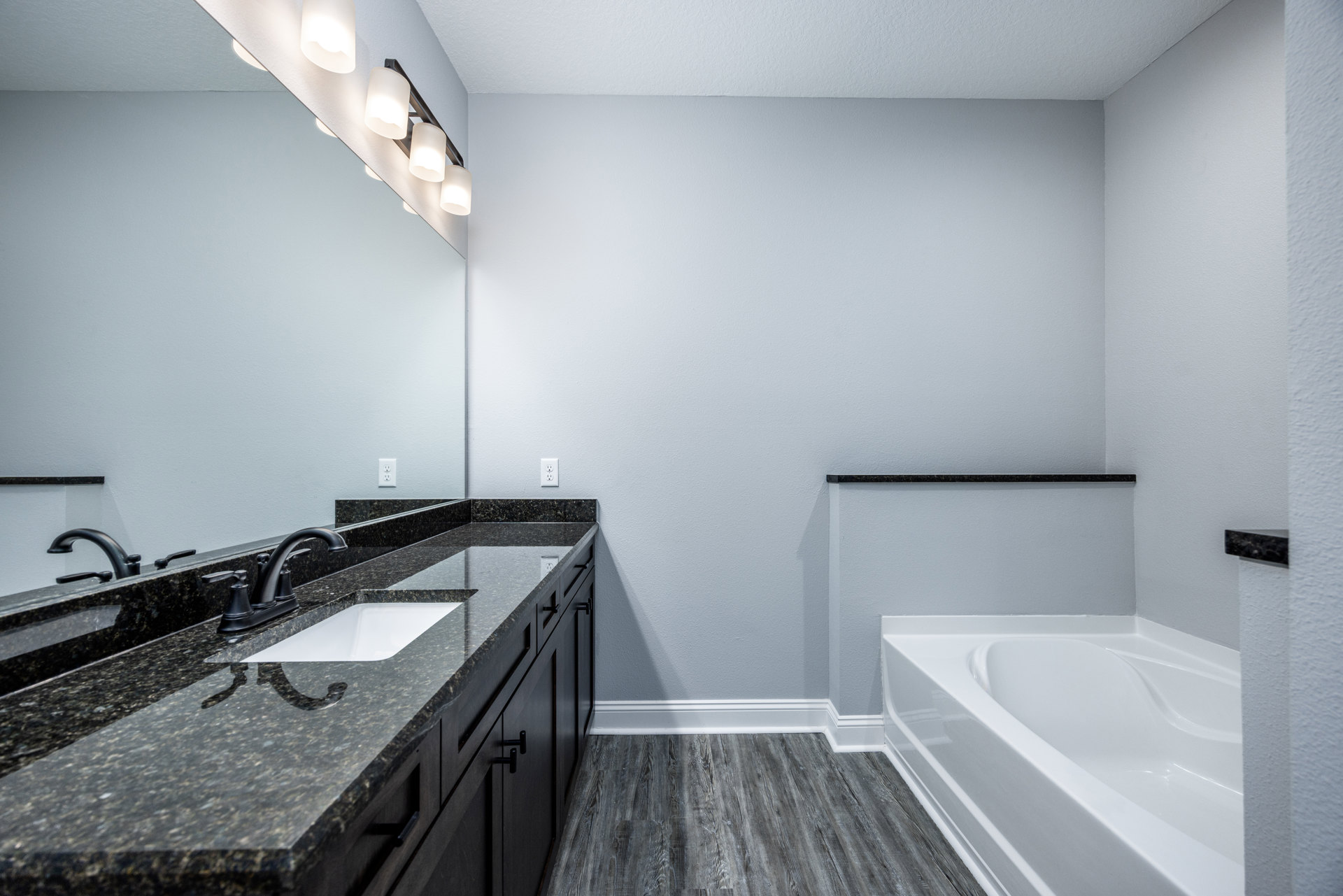 White freestanding bathtub with curved edge beside a black countertop vanity and white sink, wall-mounted faucet, large mirror, light gray tile walls and floor.