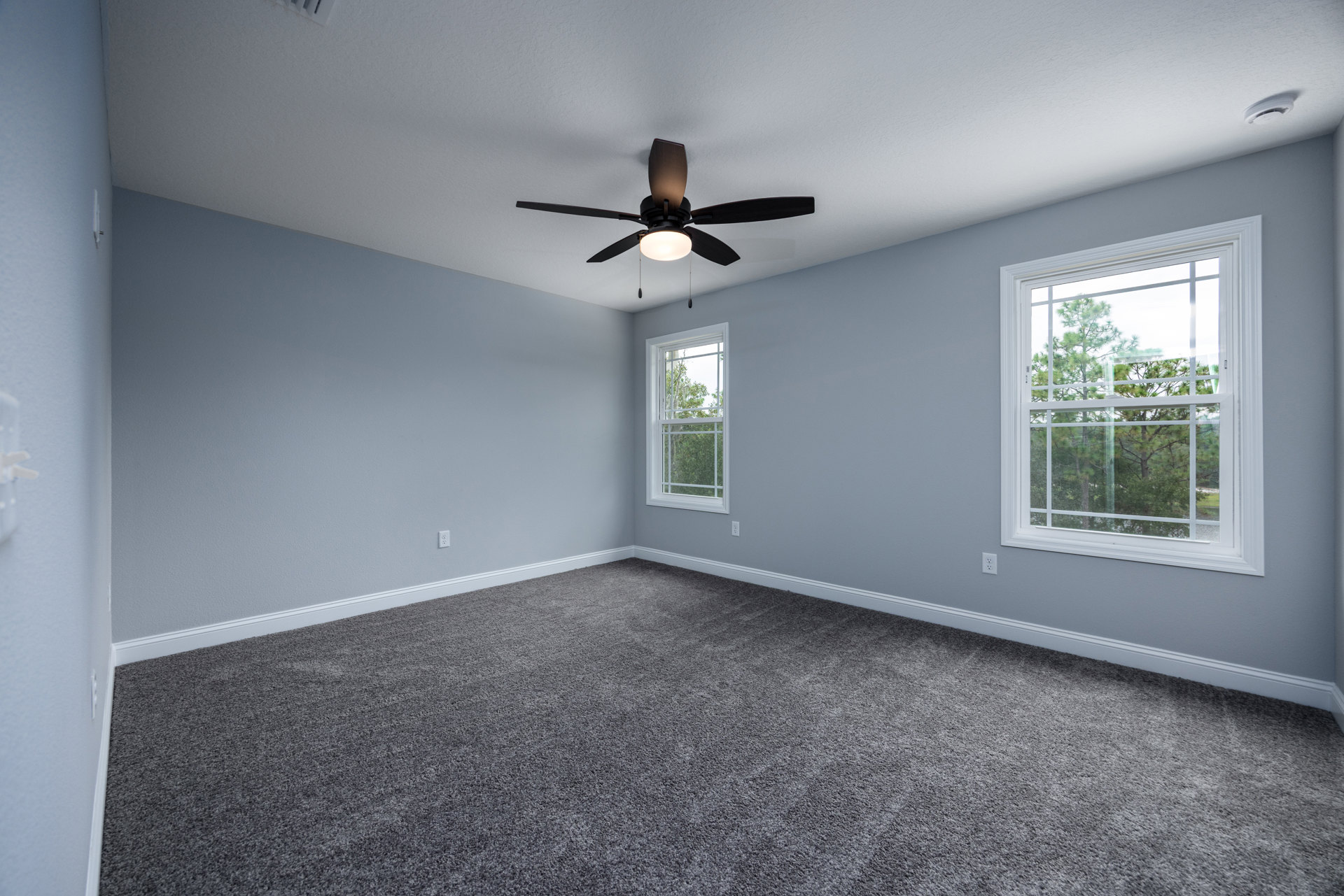 Neutral-toned room with grey carpet, white walls, ceiling fan with light, large windows showing trees outside, and smoke detector on ceiling