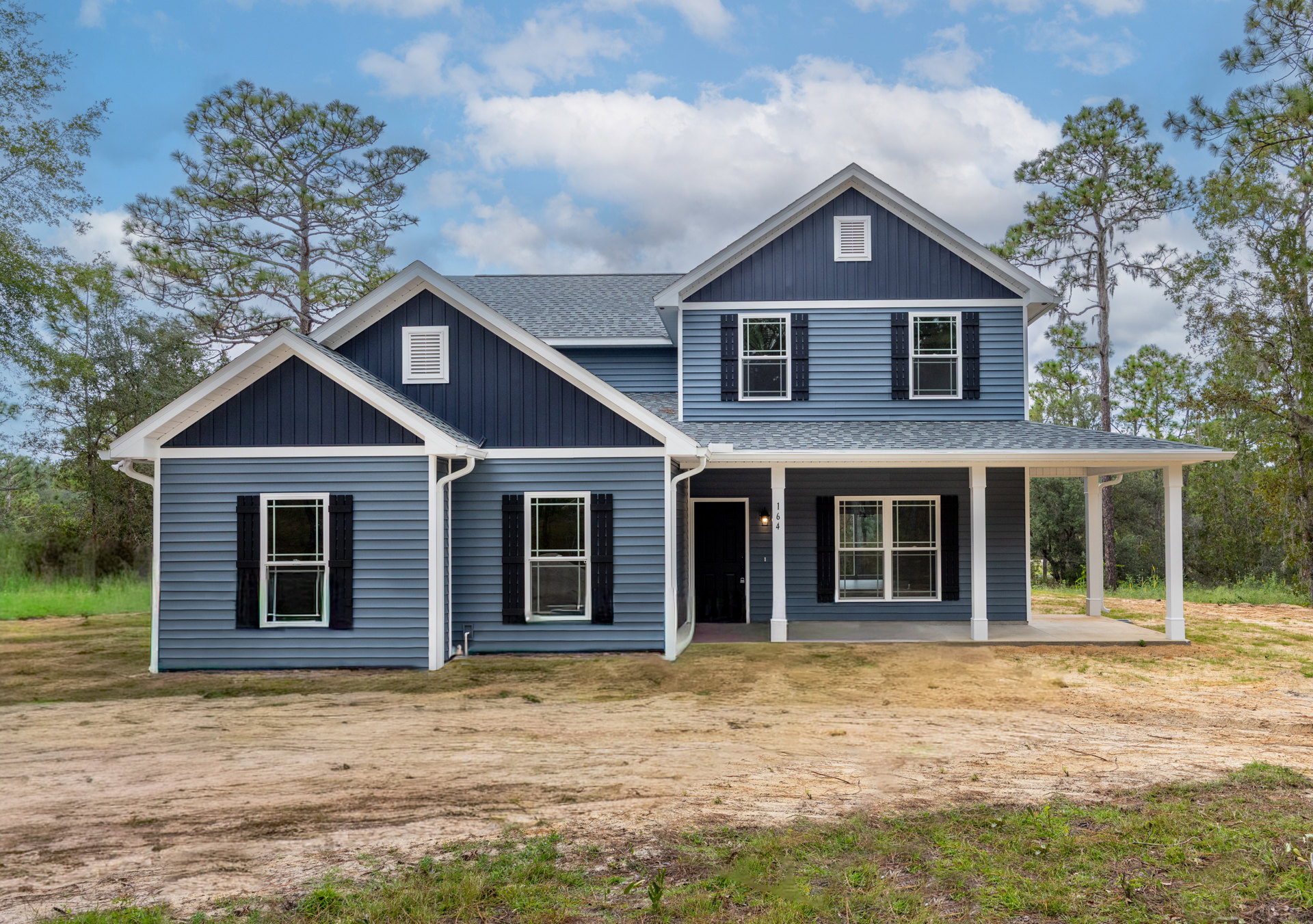 Blue siding house with white trim windows, white vent on wall, dirt yard, concrete driveway, mature trees in background, cloudy sky overhead