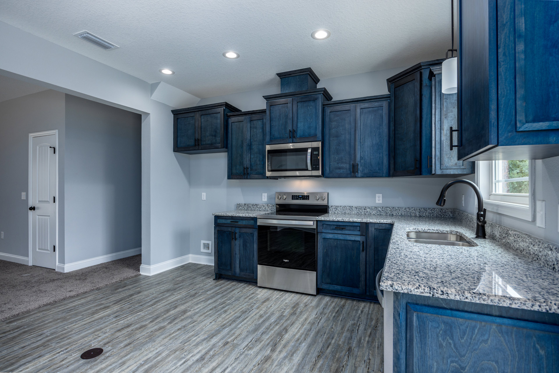 Blue kitchen cabinets with granite countertops, stainless steel microwave with open door, black glass stove, white door with black knob, close-up of undermount sink, wood flooring