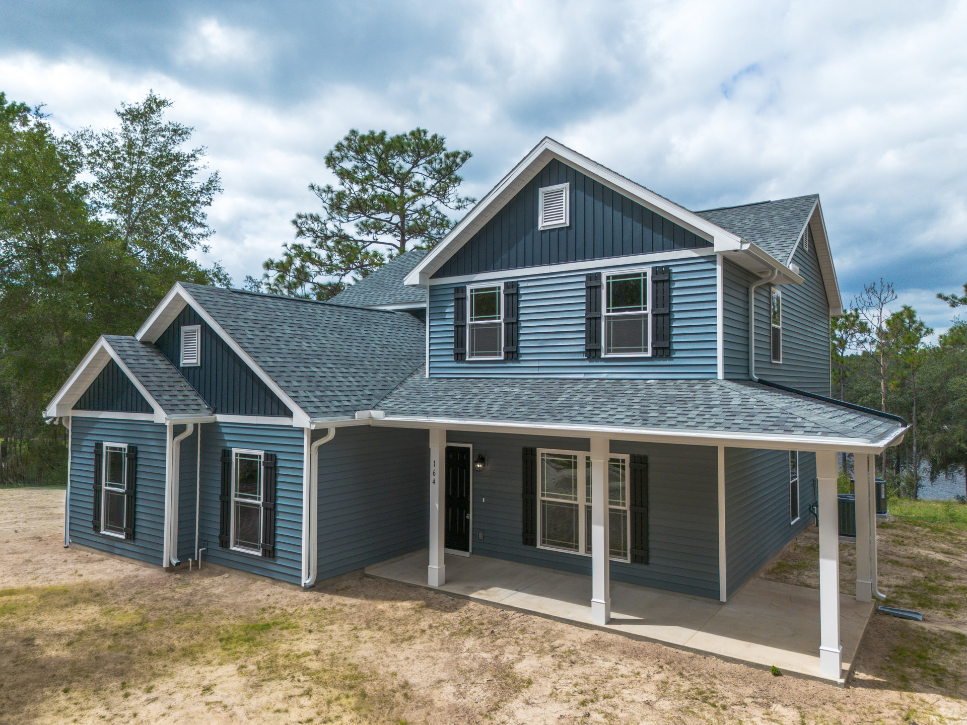 Blue siding house with spacious covered porch, white framed windows, white wall vent, and mature tree in the background under partly cloudy sky.