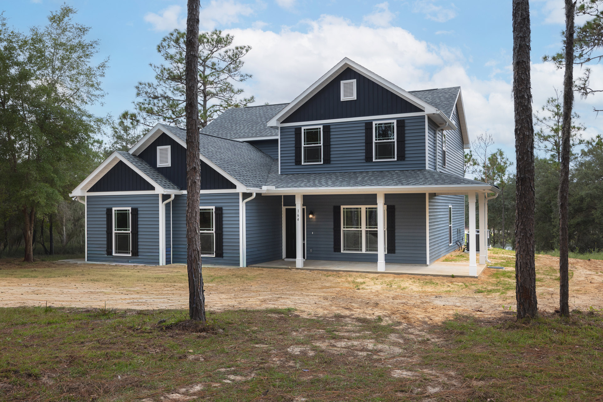 Two-story home with white-trimmed windows, covered porch, and mature tree in front yard, dirt patch near entry, cloudy sky overhead