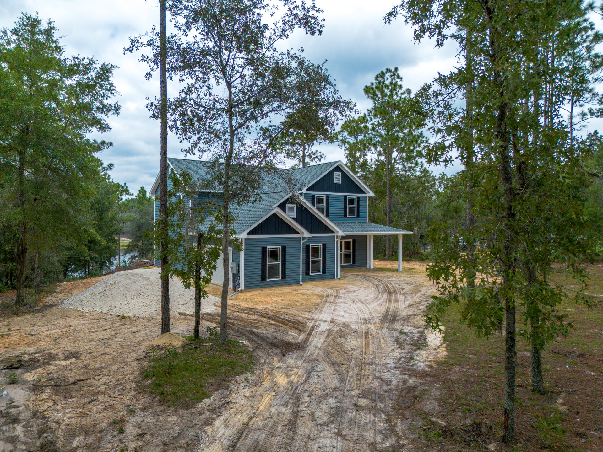White cottage-style house surrounded by mature leafy trees, grassy lawn, and dirt driveway under partly cloudy sky