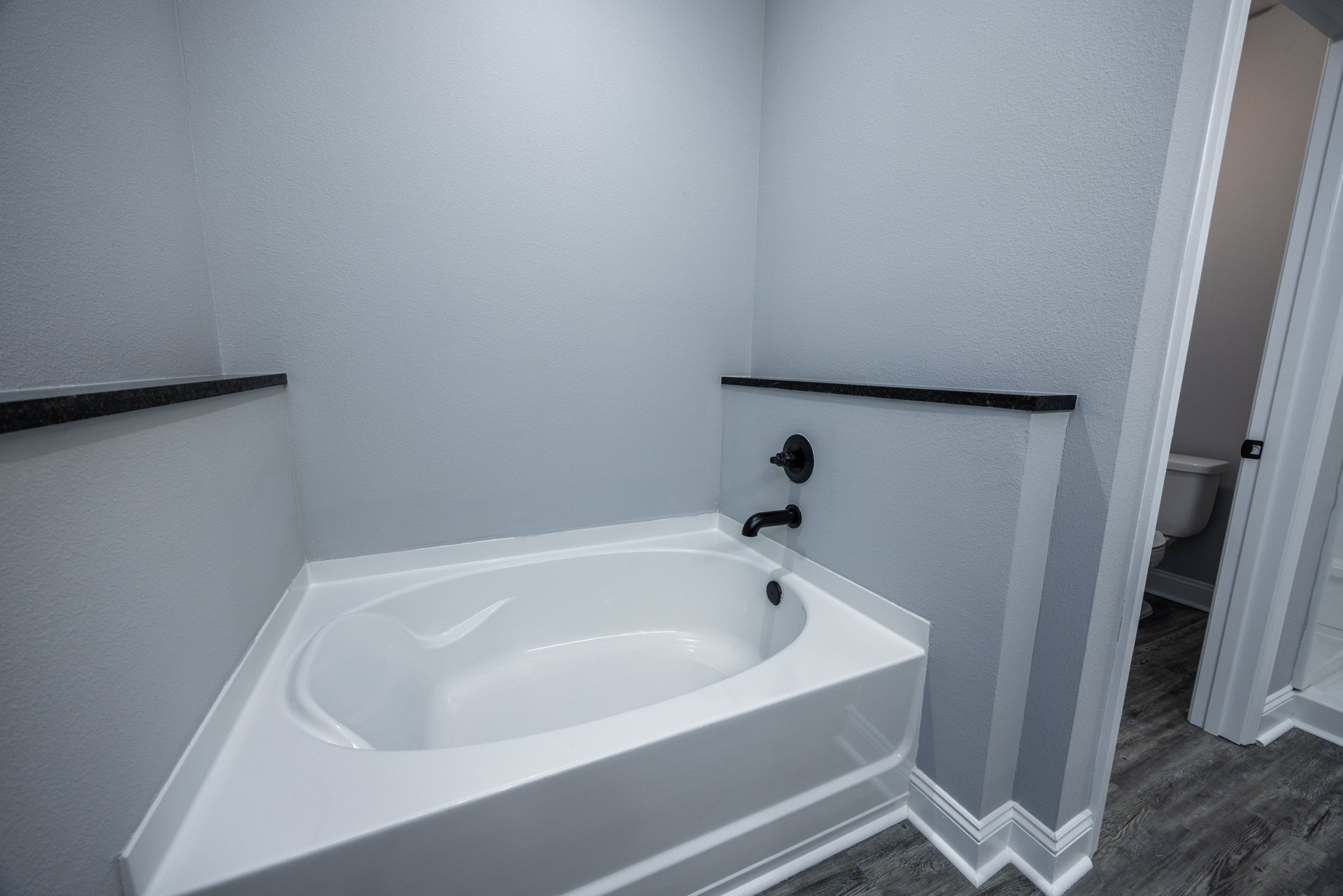 Freestanding white bathtub with curved edge, matte black wall-mounted faucet and switch, white tile walls, partial view of toilet