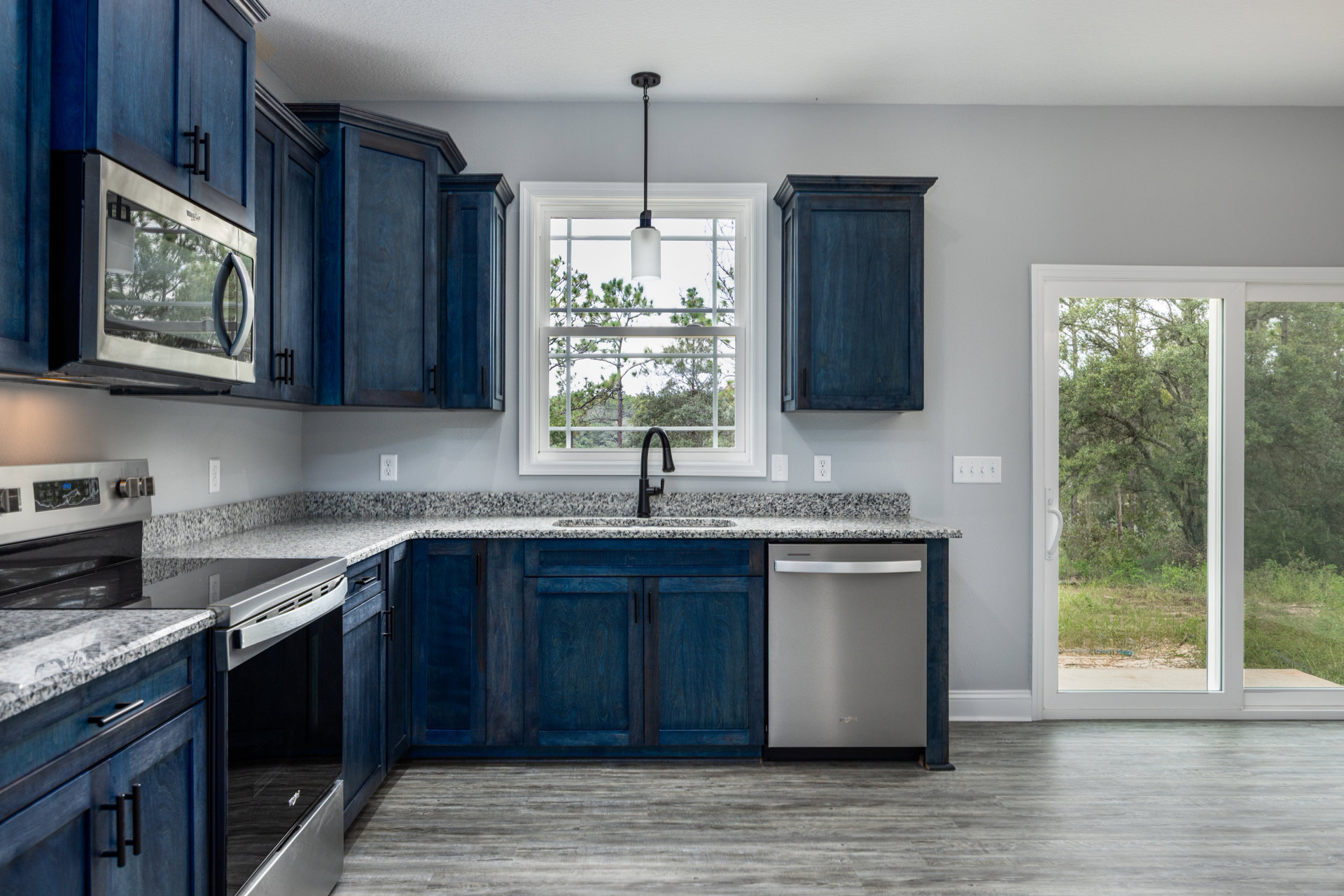 Blue kitchen cabinets with black hardware, white countertop, stainless steel refrigerator, black faucet at sink beneath window, blue door with black handle, trees visible outside.