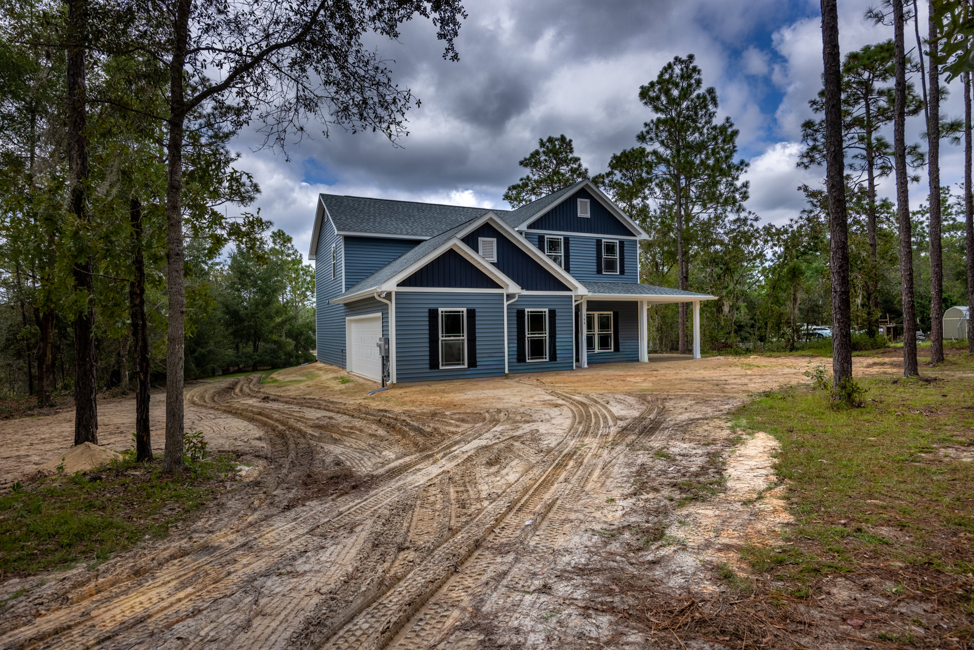 Blue house with white trim surrounded by trees, white-framed window, white door with black sign, dirt road with visible tire tracks in foreground