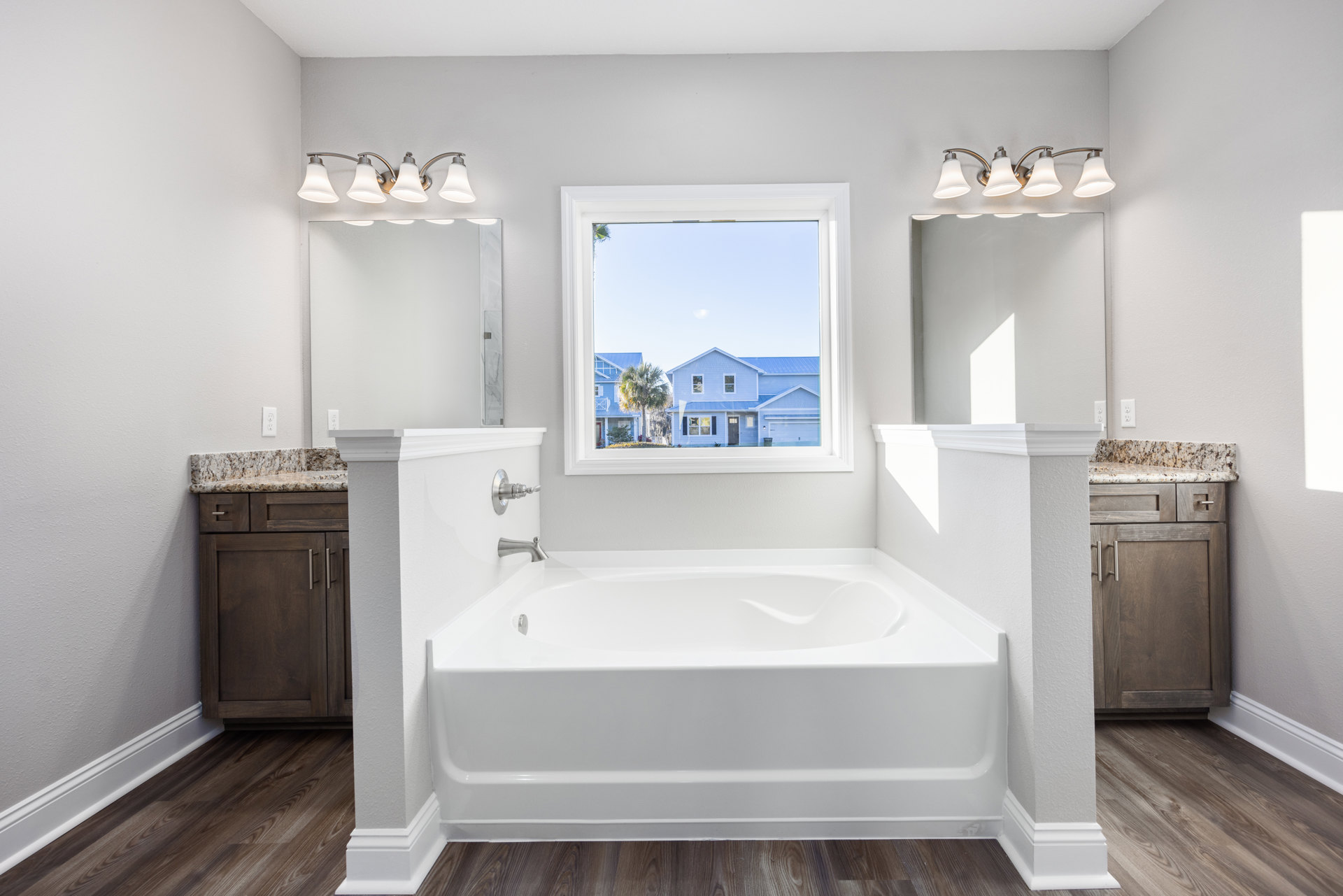 Freestanding white bathtub on gray tile floor beside a window with white trim, wooden vanity with silver handles, double light fixture above mirror