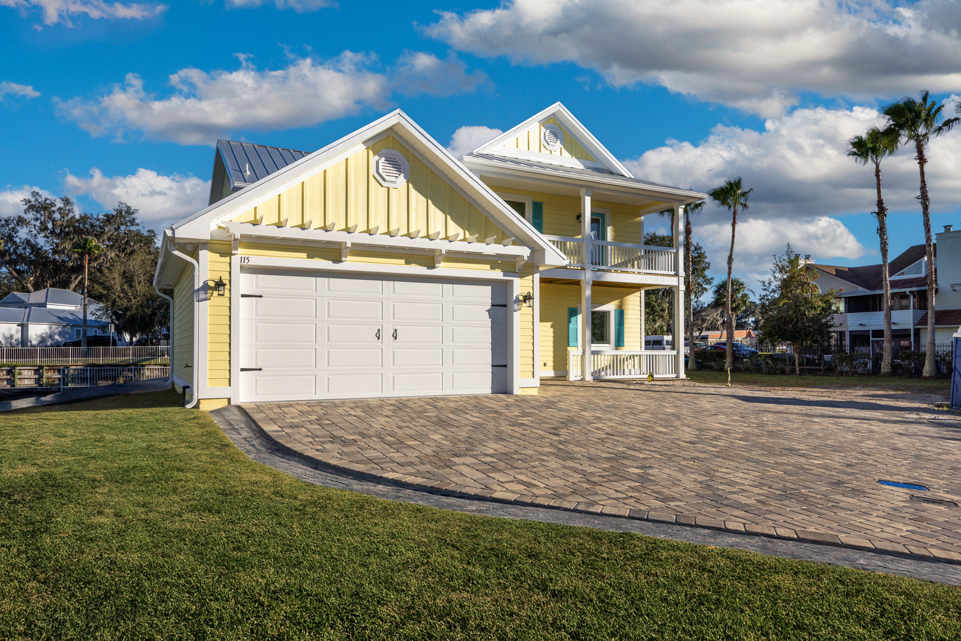 Yellow house with white trim, two-car garage, white vent on exterior wall, palm trees in front, brick driveway bordered by grass, cloudy sky overhead.