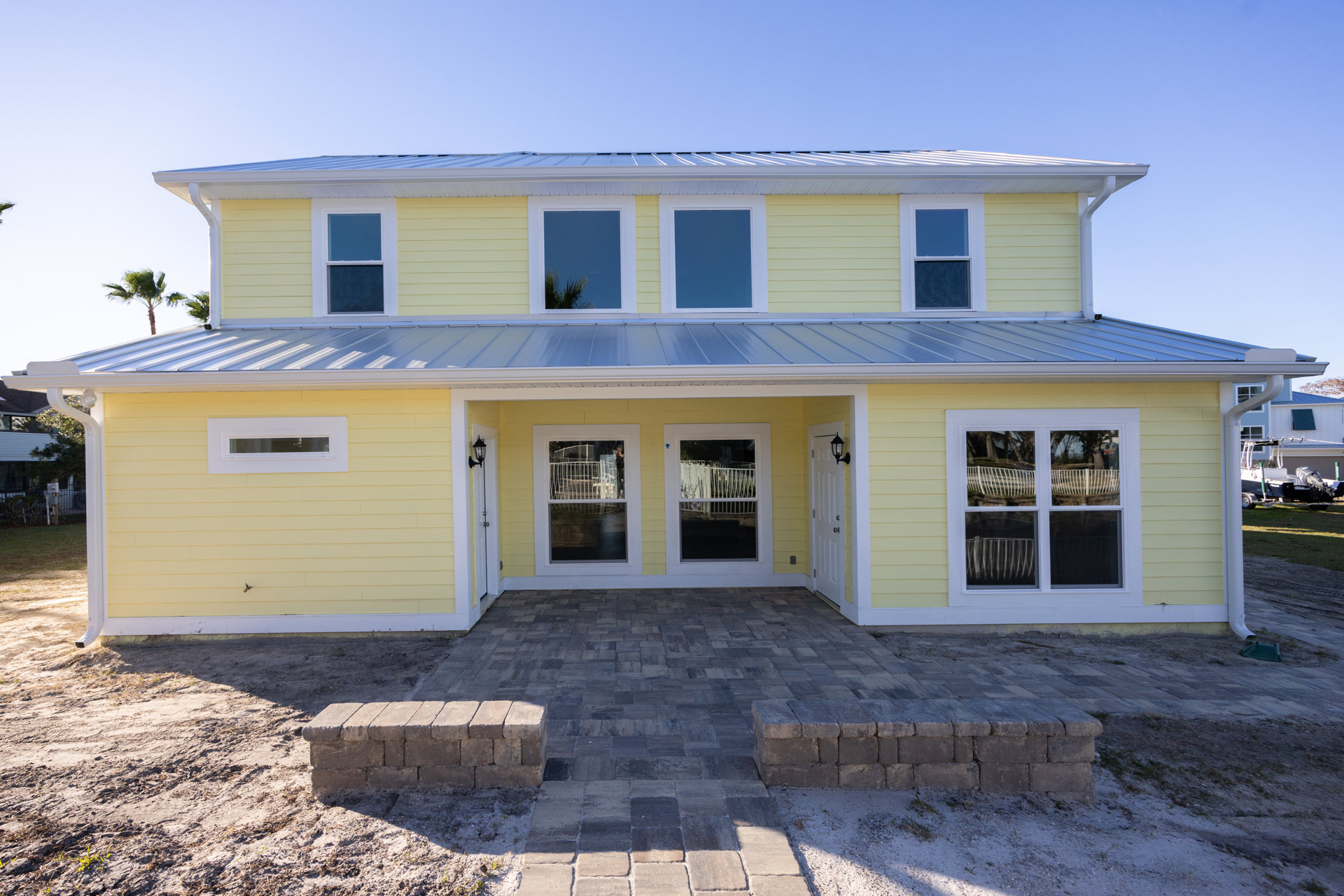 Yellow siding house with metal roof, multiple windows, brick patio, white railing, palm tree, and blue sky.