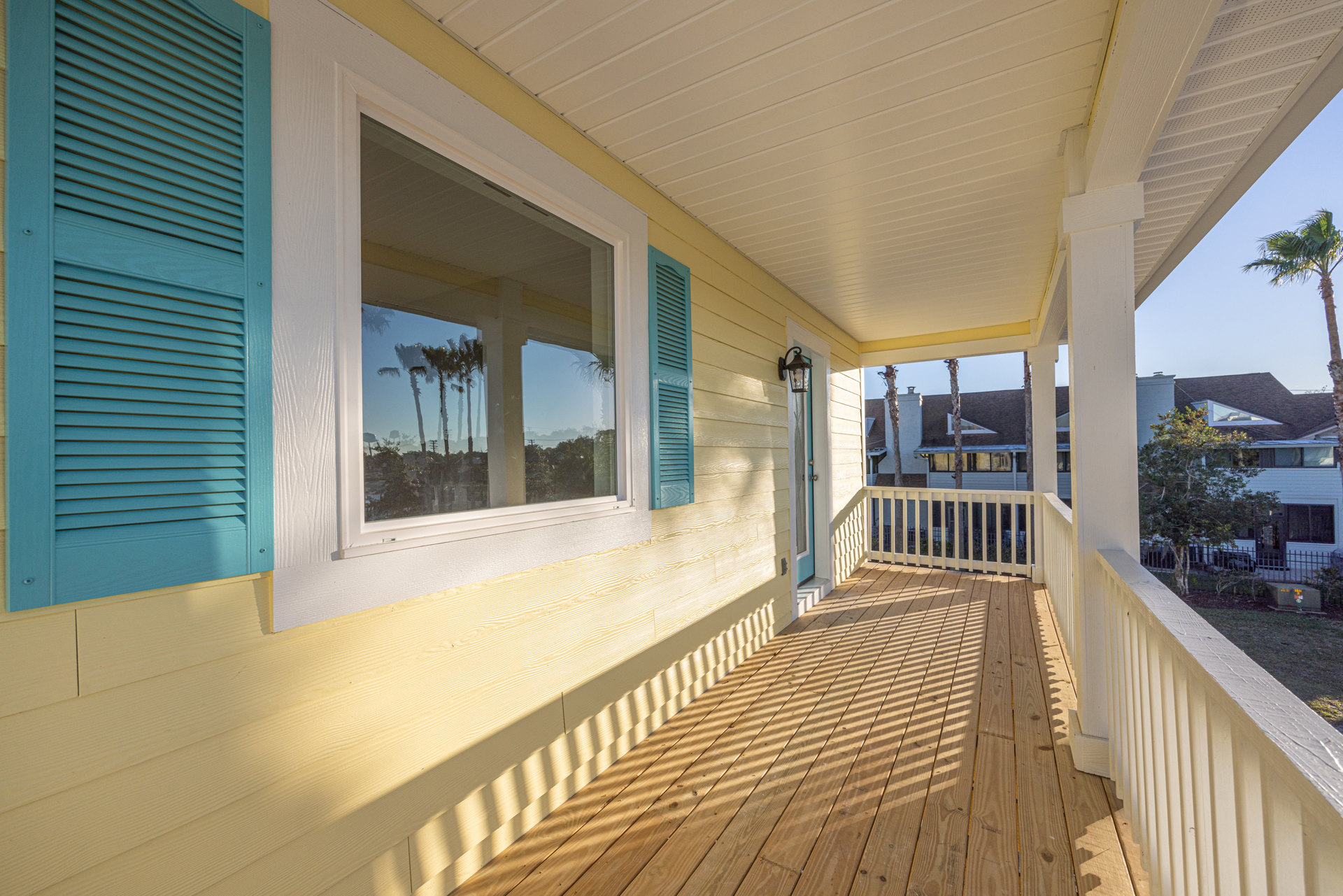 Wood deck porch with white-trimmed windows, blue shutters, and palm trees reflected in glass under clear sky