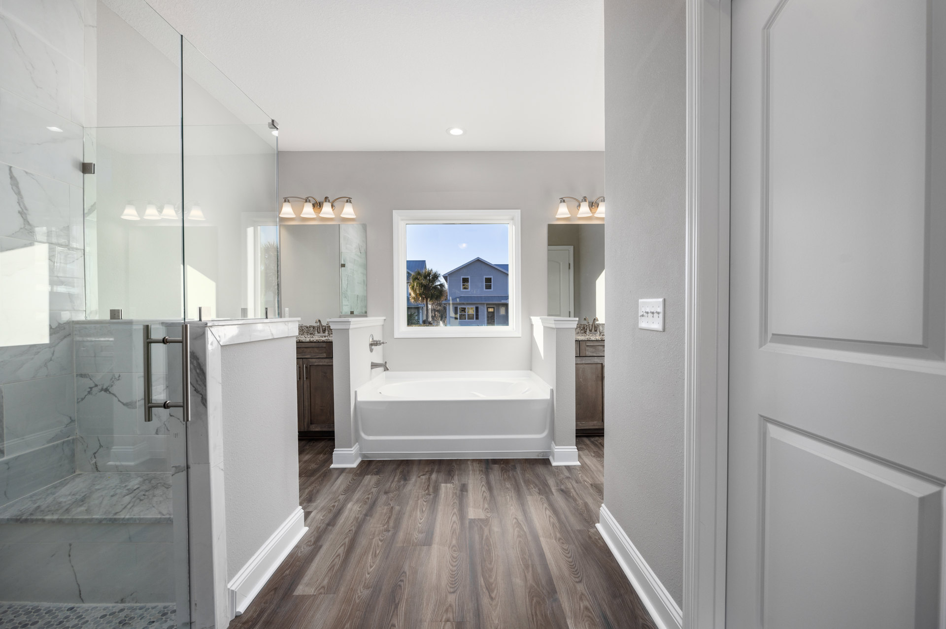 Freestanding white bathtub beside glass-enclosed shower, white walls with metal hooks, wide window overlooking palm tree and neighboring house, light wood flooring throughout