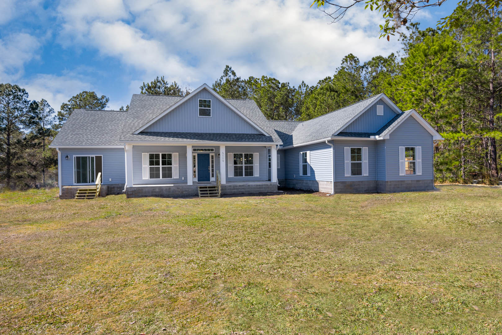 White cottage-style home with blue door and white trim, surrounded by green lawn and mature trees, cloudy sky overhead