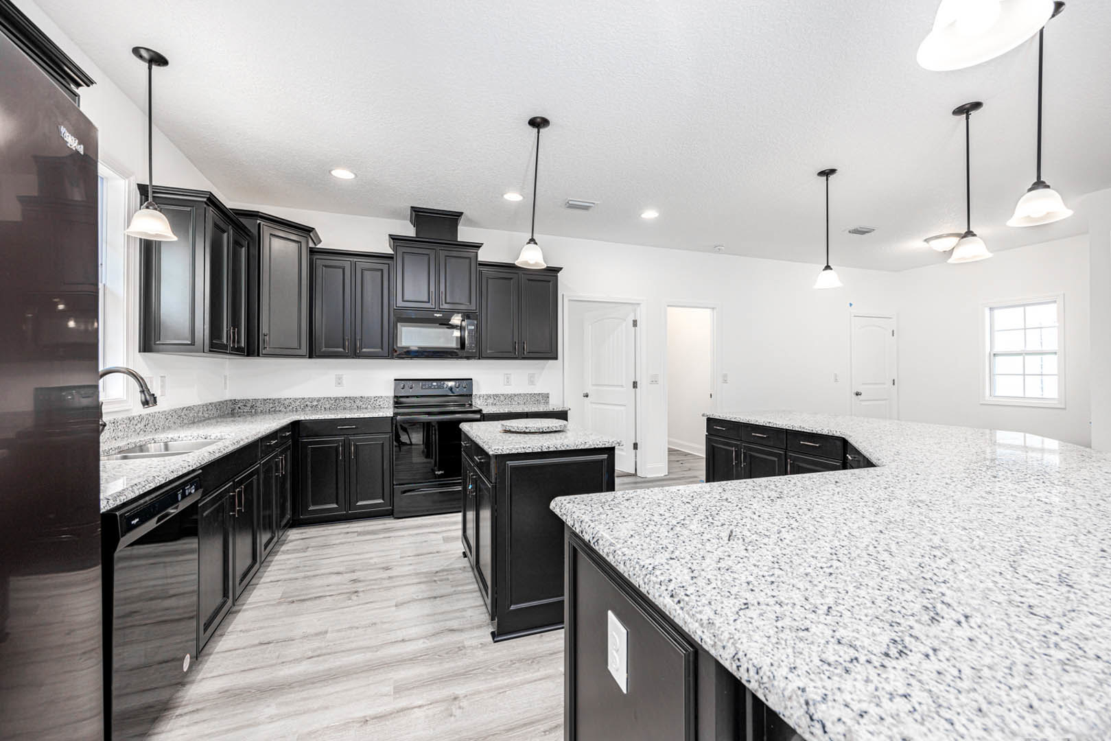 Black kitchen cabinets paired with white countertops, stainless steel sink, black speckled marble island, modern pendant lighting, light wood flooring, large window with natural