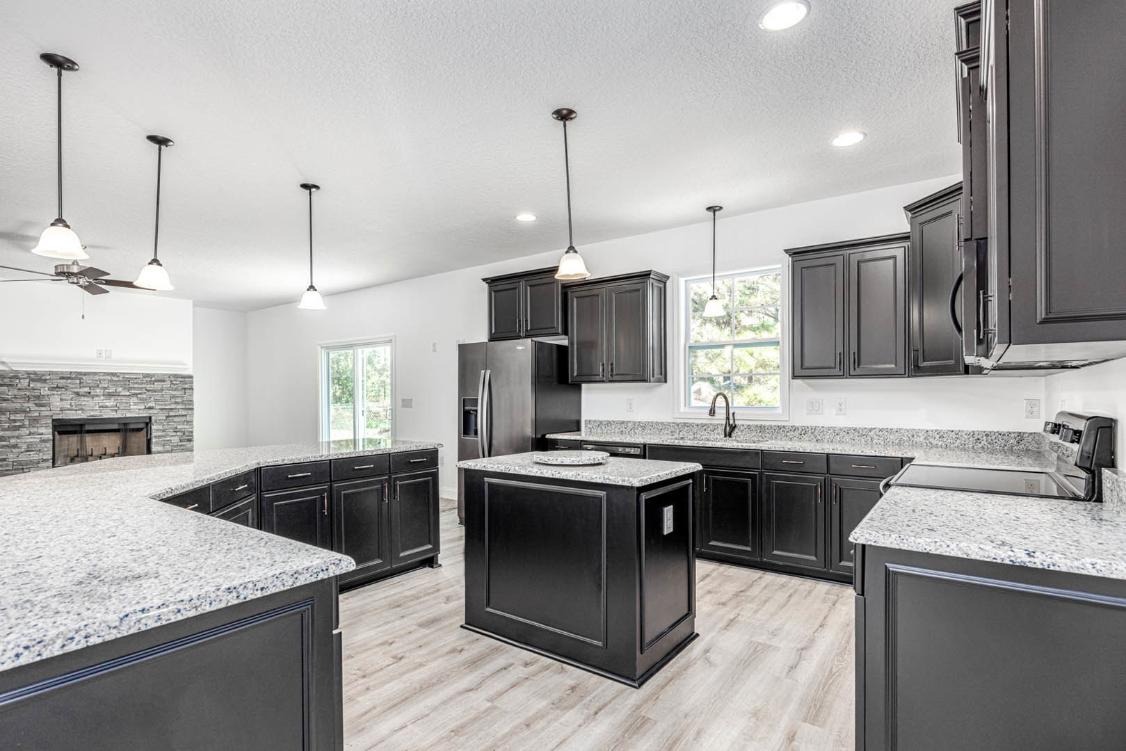 Black kitchen cabinets with granite countertops, white kitchen island, stainless steel appliances, stone fireplace wall, large window, and modern sink fixtures.