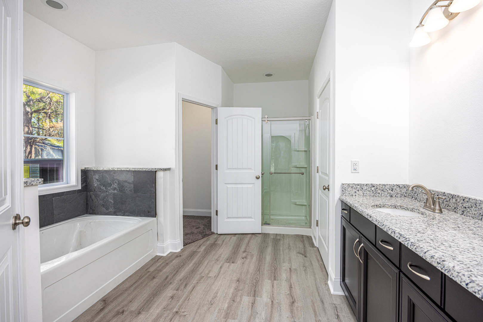 Bathroom featuring a freestanding white bathtub on wood flooring, glass shower enclosure with metal bar, granite countertop with sink and faucet, white door with silver knob